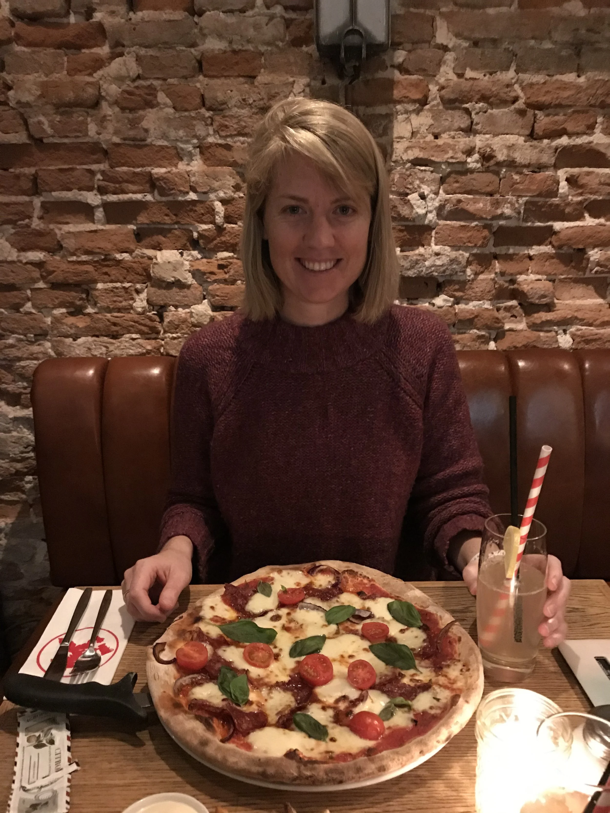 A woman with blonde hair and a maroon sweater sitting at a restaurant table with a pizza topped with cherry tomatoes, basil, and mozzarella, and a glass of a lemon drink with a straw.
