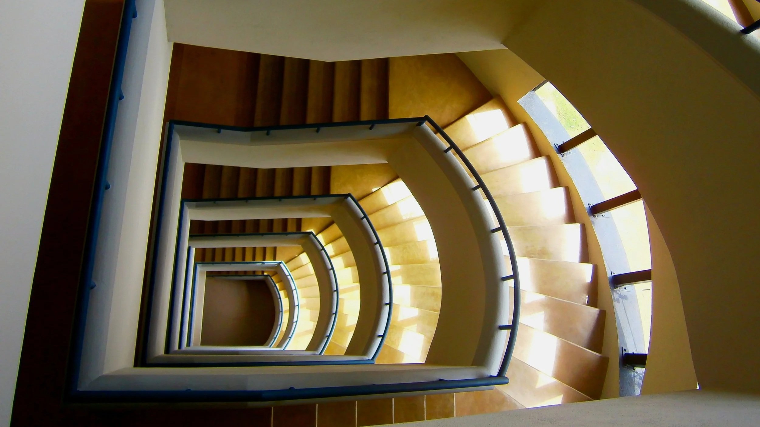 A view looking down a spiral staircase with wooden steps, white railings, and black handrails, with large windows allowing natural light to illuminate the staircase.