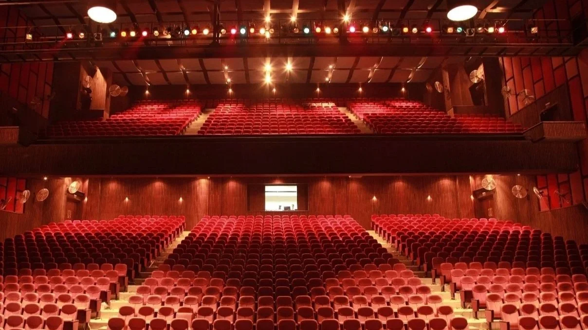 Empty theater with red seats, wooden walls, and stage lighting, viewed from the audience area.