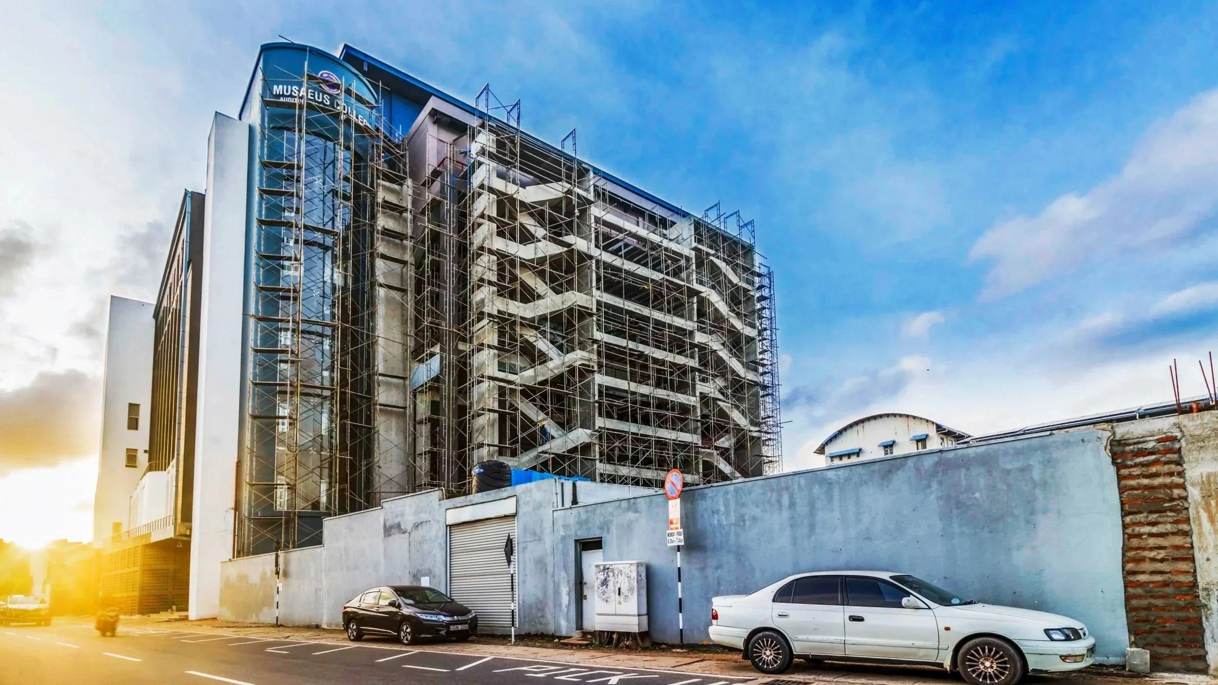 A high-rise building under construction with scaffolding surrounding it, situated near a parking lot with two cars, and a blue sky with some clouds in the background.