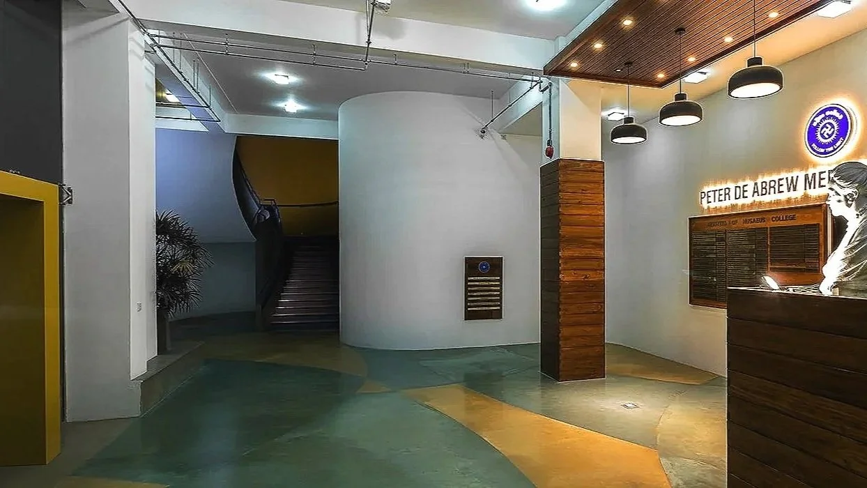 Modern college lobby with wooden accents, a reception desk, and college name sign, illuminated by ceiling lights.