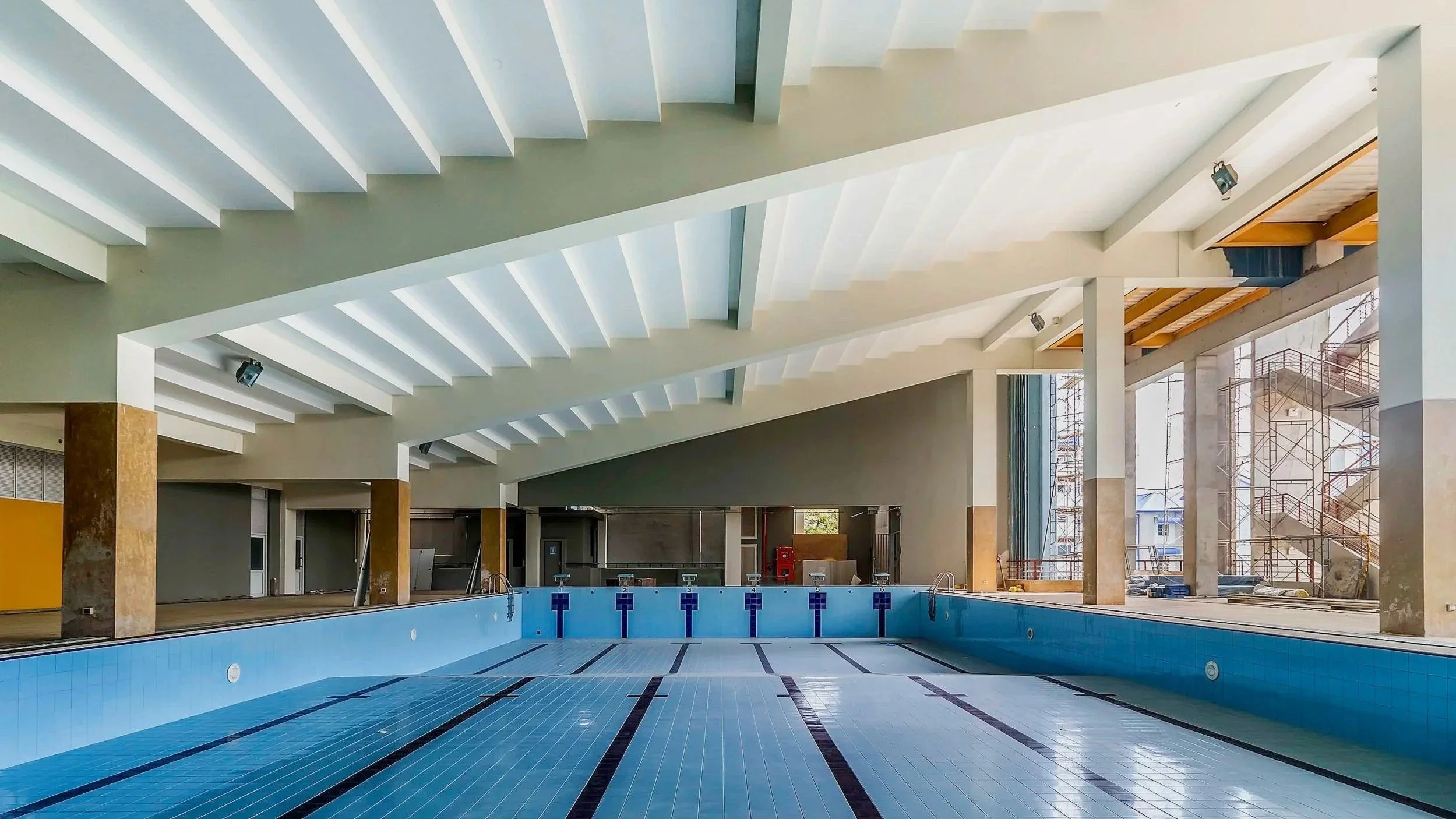 Indoor swimming pool area with blue tiles, empty pool, and modern architecture including white ceiling beams and large windows.