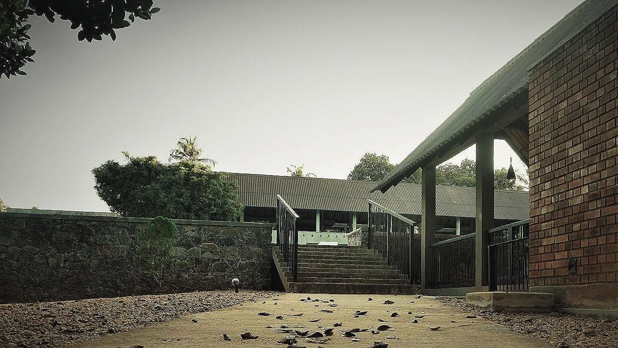 An outdoor scene showing concrete stairs with black metal railings, leading up to a building with brick and wooden exterior, surrounded by trees and a stone wall.