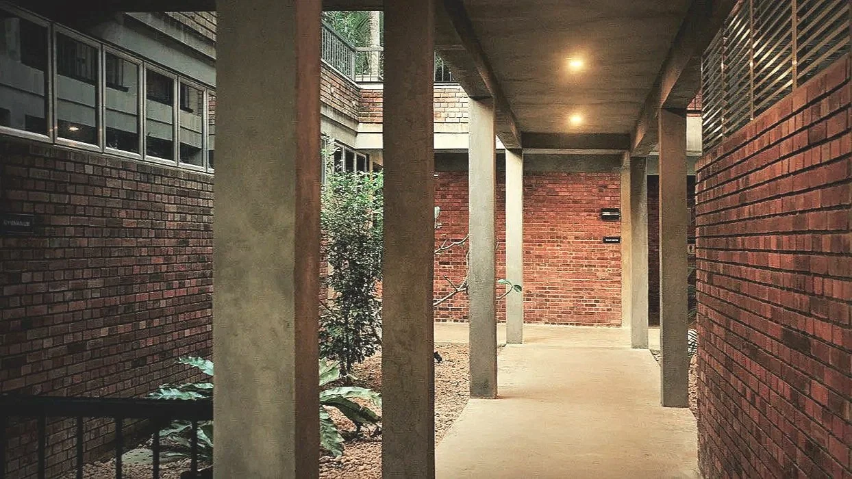 A covered outdoor walkway with brick walls, concrete pillars, and a concrete ceiling, with small plants along the side and a building with windows in the background.