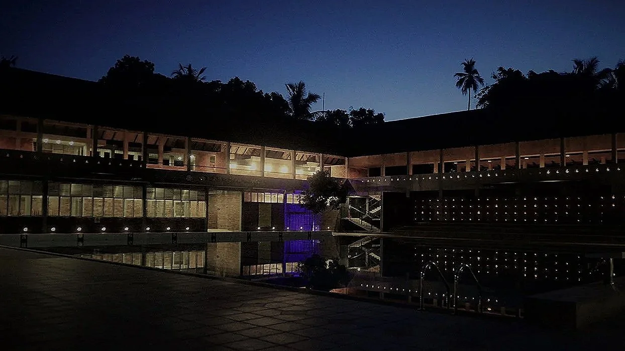 An outdoor swimming pool at night, surrounded by a building with large windows and a lit interior, with palm trees silhouetted against a dark sky.