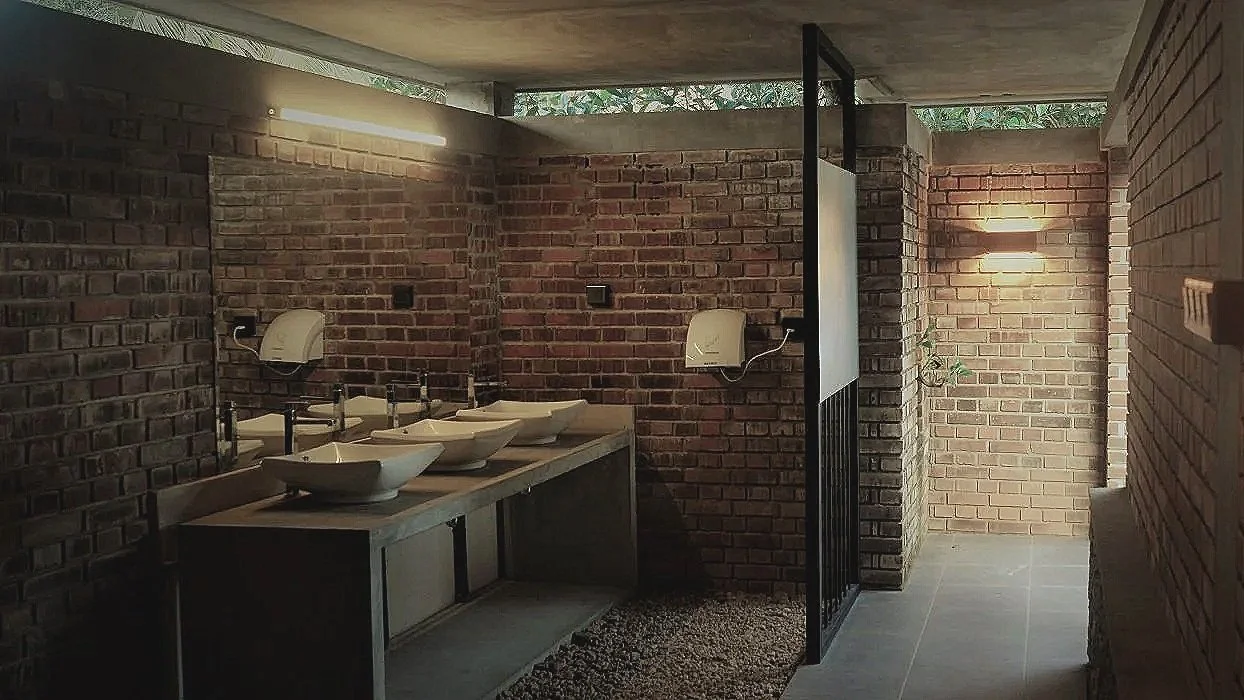 Public restroom with four sinks and soap dispensers, brick walls, modern lighting, and a semi-open roof letting in natural light.