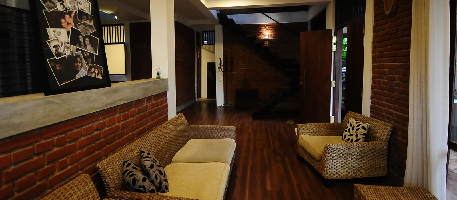Interior of a cozy living room with brick walls, wood flooring, wicker furniture with cushions and patterned throw pillows, a gallery wall with photographs, and an open door leading outside.