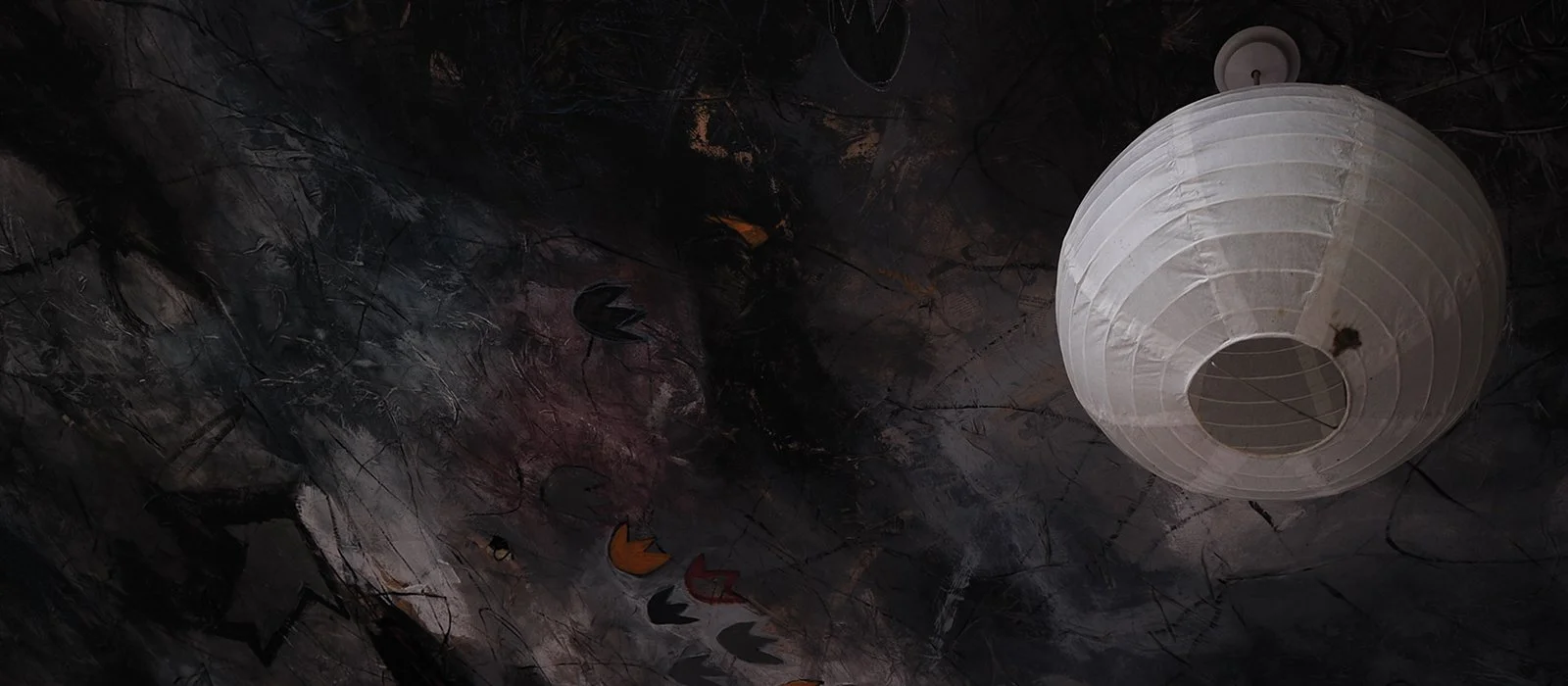 A large white paper lantern hanging from a ceiling with a dark, textured background, possibly painted or decorated with abstract patterns.