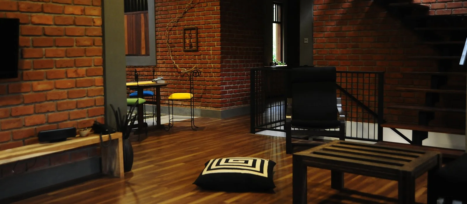 Interior of a modern home with exposed brick walls, wooden floors, and black furniture, including a chair, a small table, and a padded cushion on the floor.