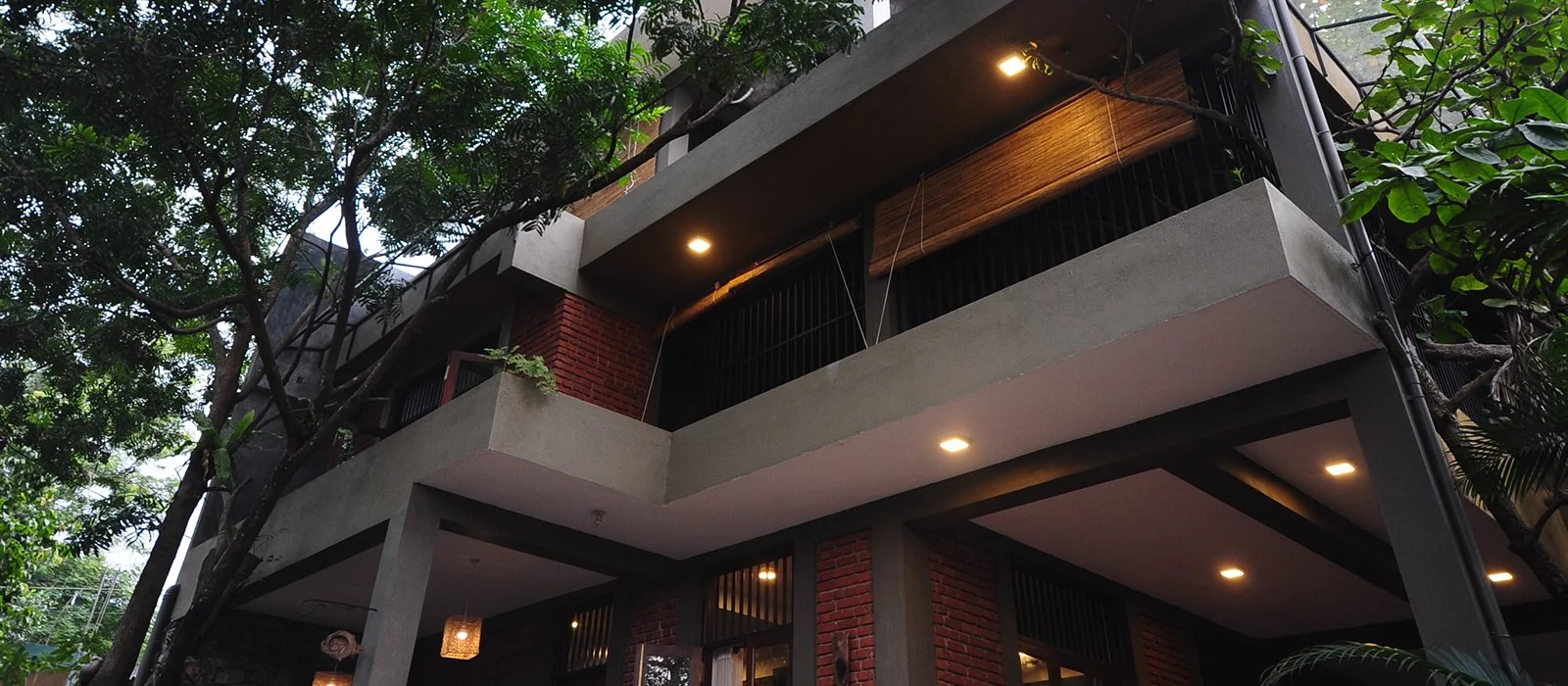 Low-angle view of a modern multi-story residential building with exposed brick walls, concrete balconies, and lush green trees surrounding it, with exterior lighting turned on.