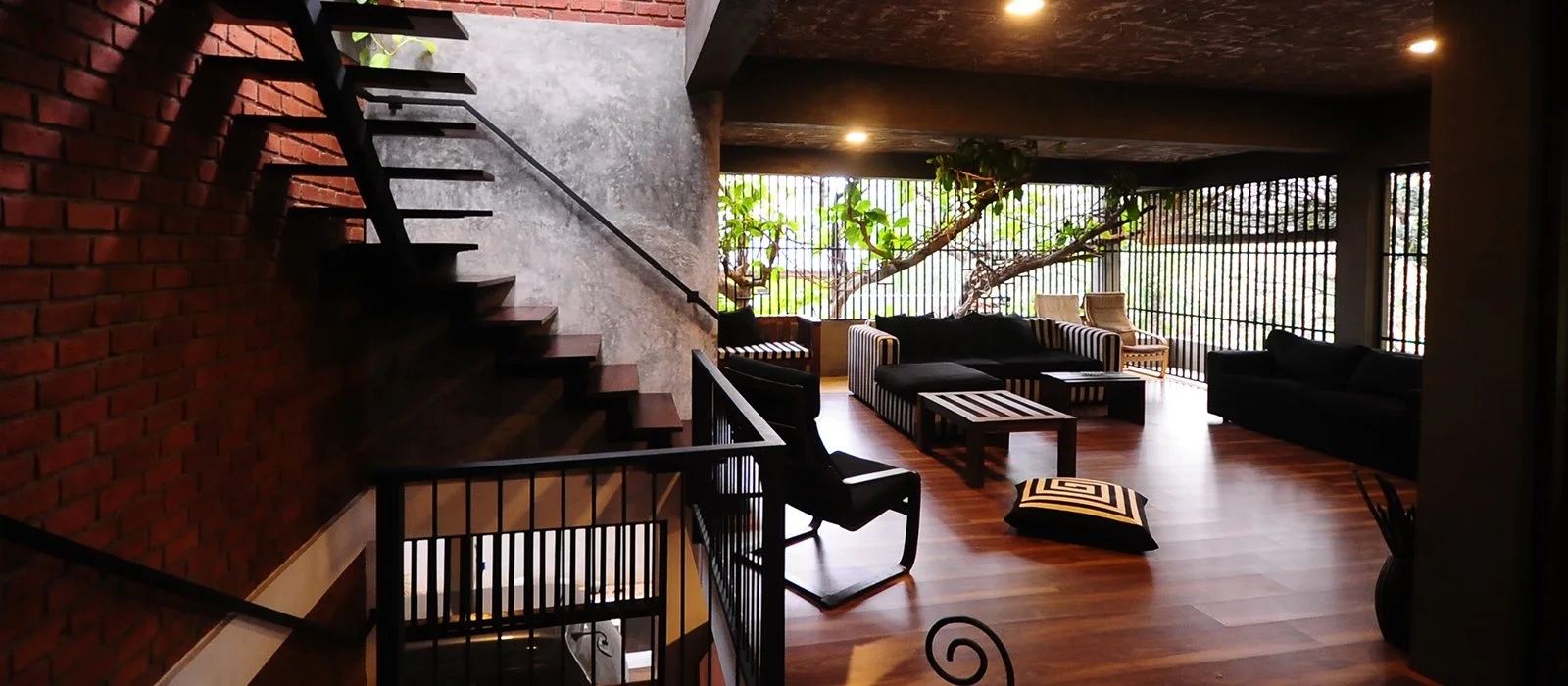 Living room with large windows, black and striped furniture, a wooden coffee table, and a staircase with black railing on the left side.