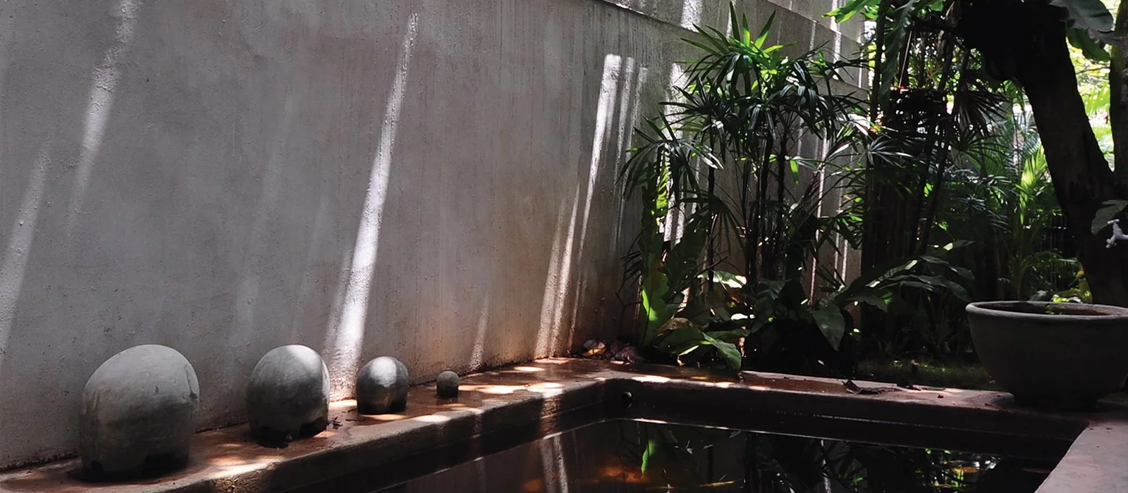Decorative outdoor fountain with five smooth, rounded stone spheres of varying sizes on a brick ledge, set against a plain concrete wall with shadows and lush green plants nearby.