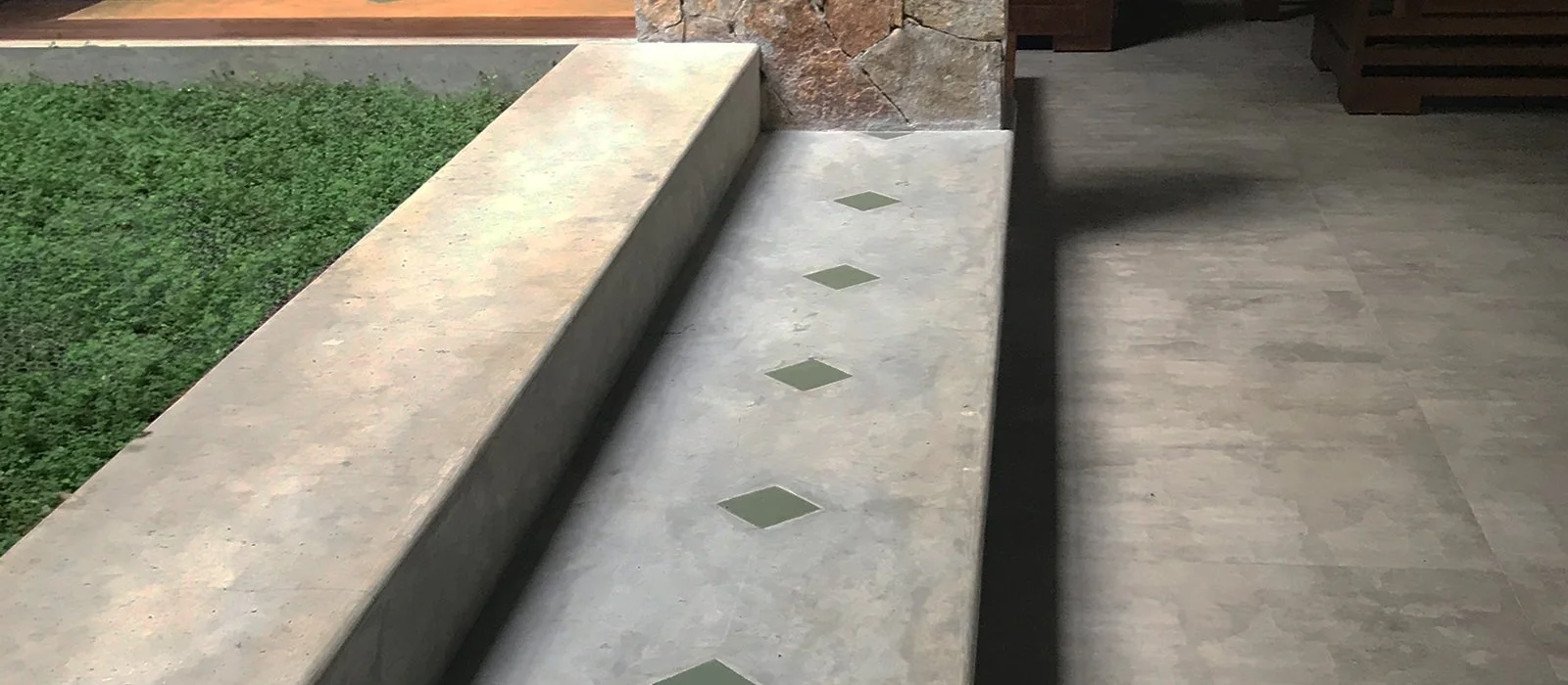 Concrete bench with small square tiles, next to a patch of green grass, against a stone wall in an indoor or outdoor setting.