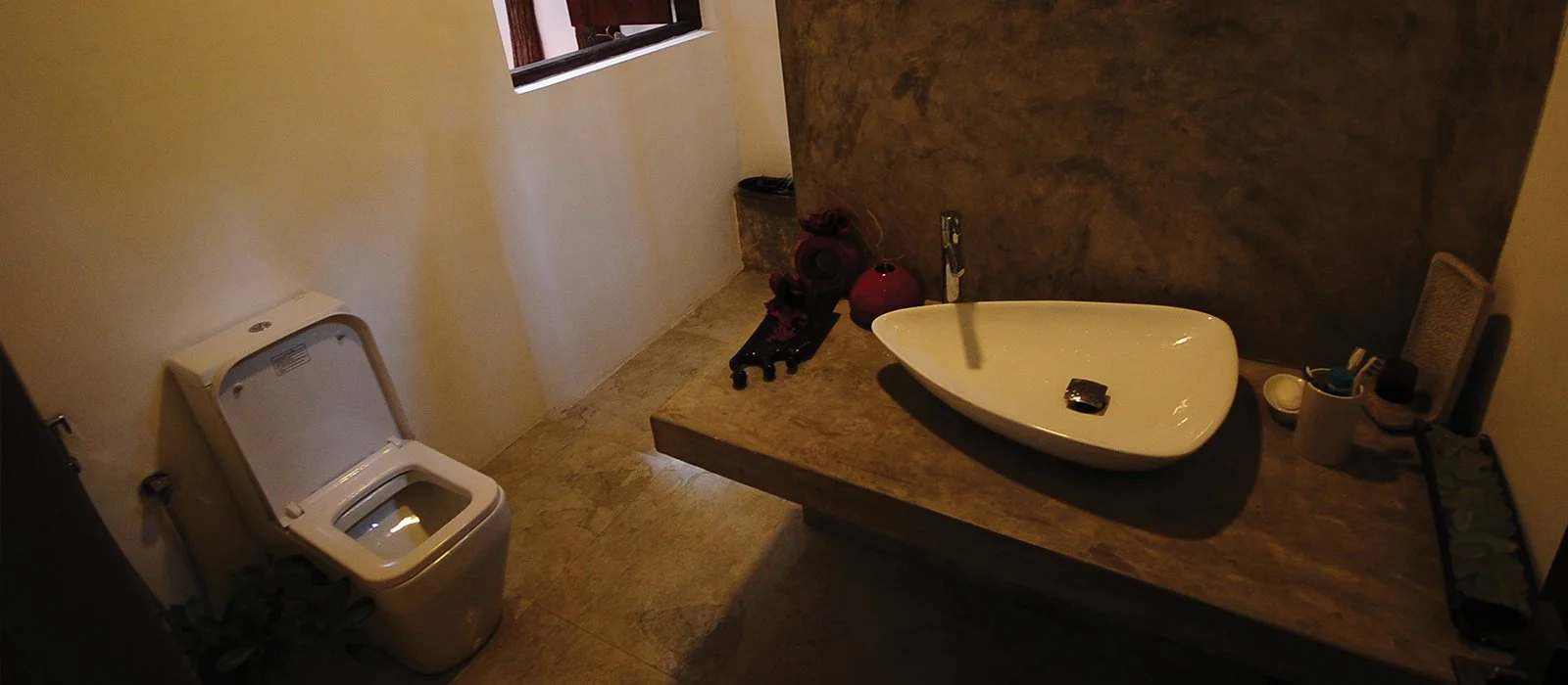 A bathroom with a white toilet on the left and a marble countertop with a vessel sink on the right. Items on the countertop include a red decorative object, a rolled towel, a soap dish, and a container with toothbrushes. There is a towel holder with