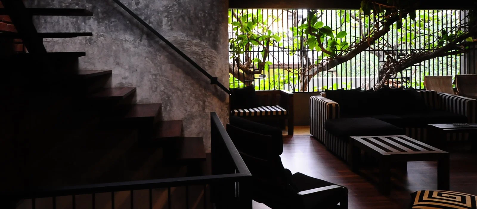 Interior of a living room with a staircase on the left, a sitting area with black and striped furniture, and a large window with greenery outside.