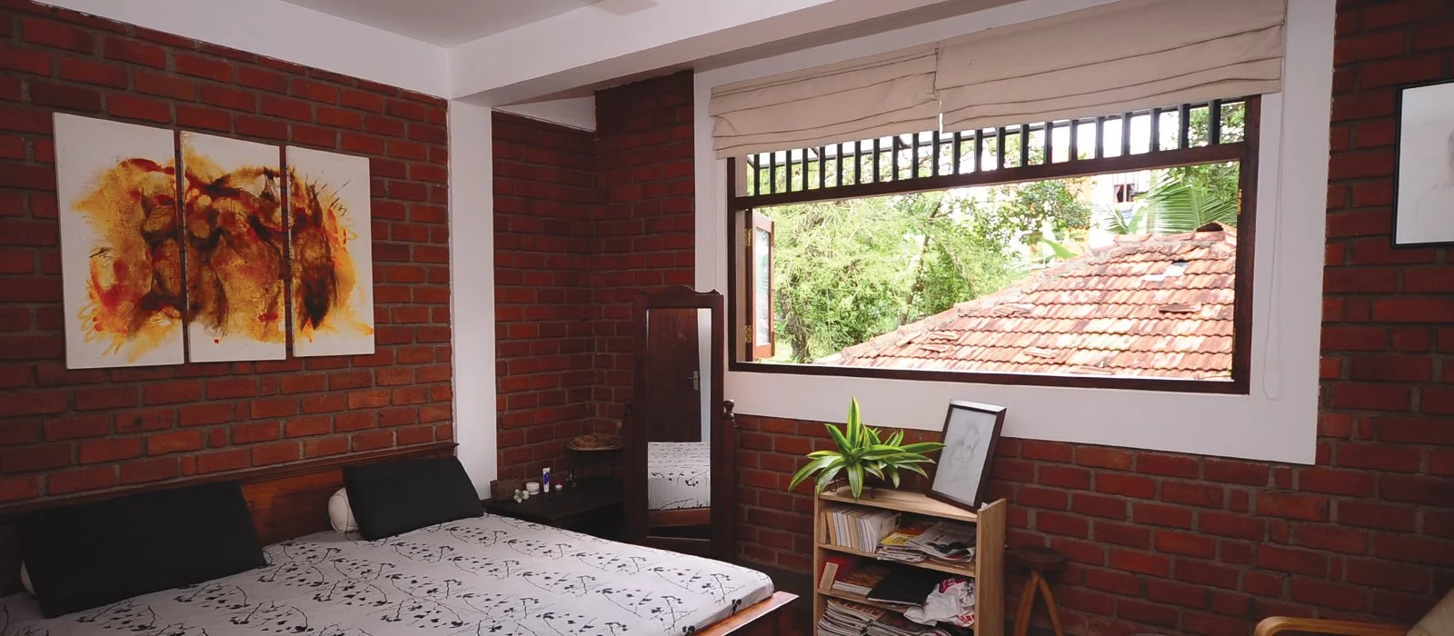A cozy bedroom with exposed brick walls, a bed with black and white bedding, a small black nightstand, wall art, an open window with a view of tropical trees and a tiled roof, and a bookshelf with a potted plant.