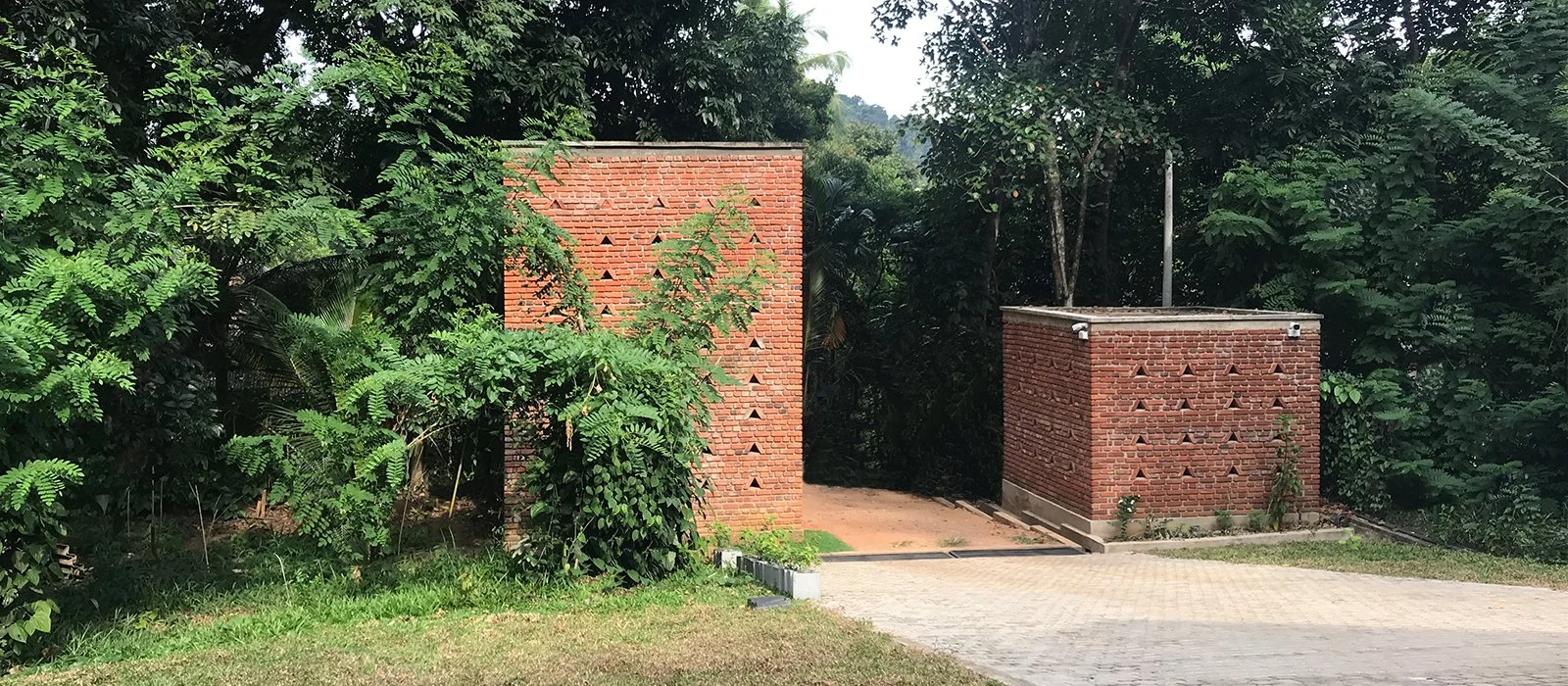 Two red brick structures with triangular holes, surrounded by green trees and plants, with a paved driveway and grass in the foreground.