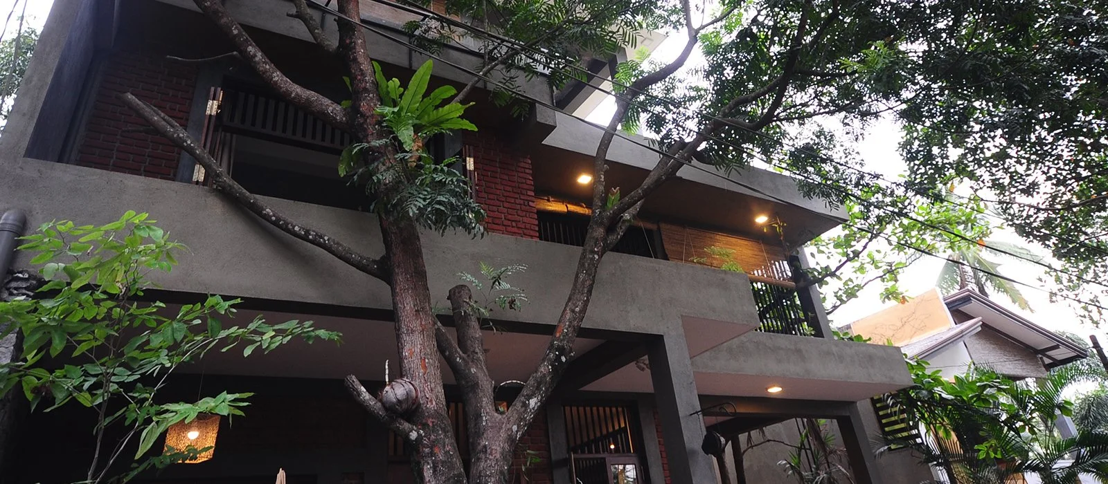 A modern multi-story house with a flat roof and concrete balconies, surrounded by trees and greenery.