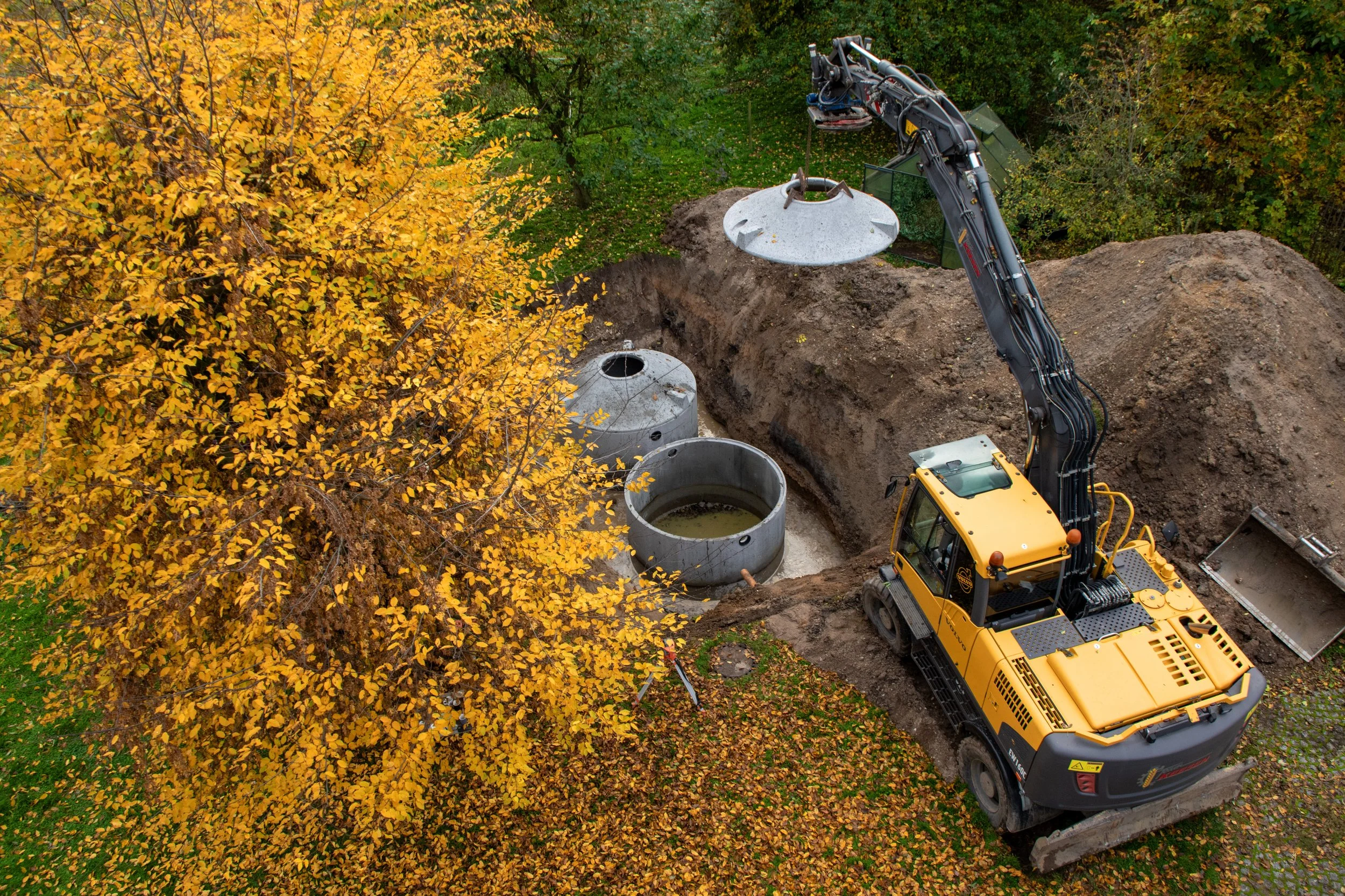 Ein Bagger arbeitet an einer Erdbaugrube, in der mehrere runde, graue Betonrohre liegen, umgeben von gelben und grünen Bäumen im Herbst.
