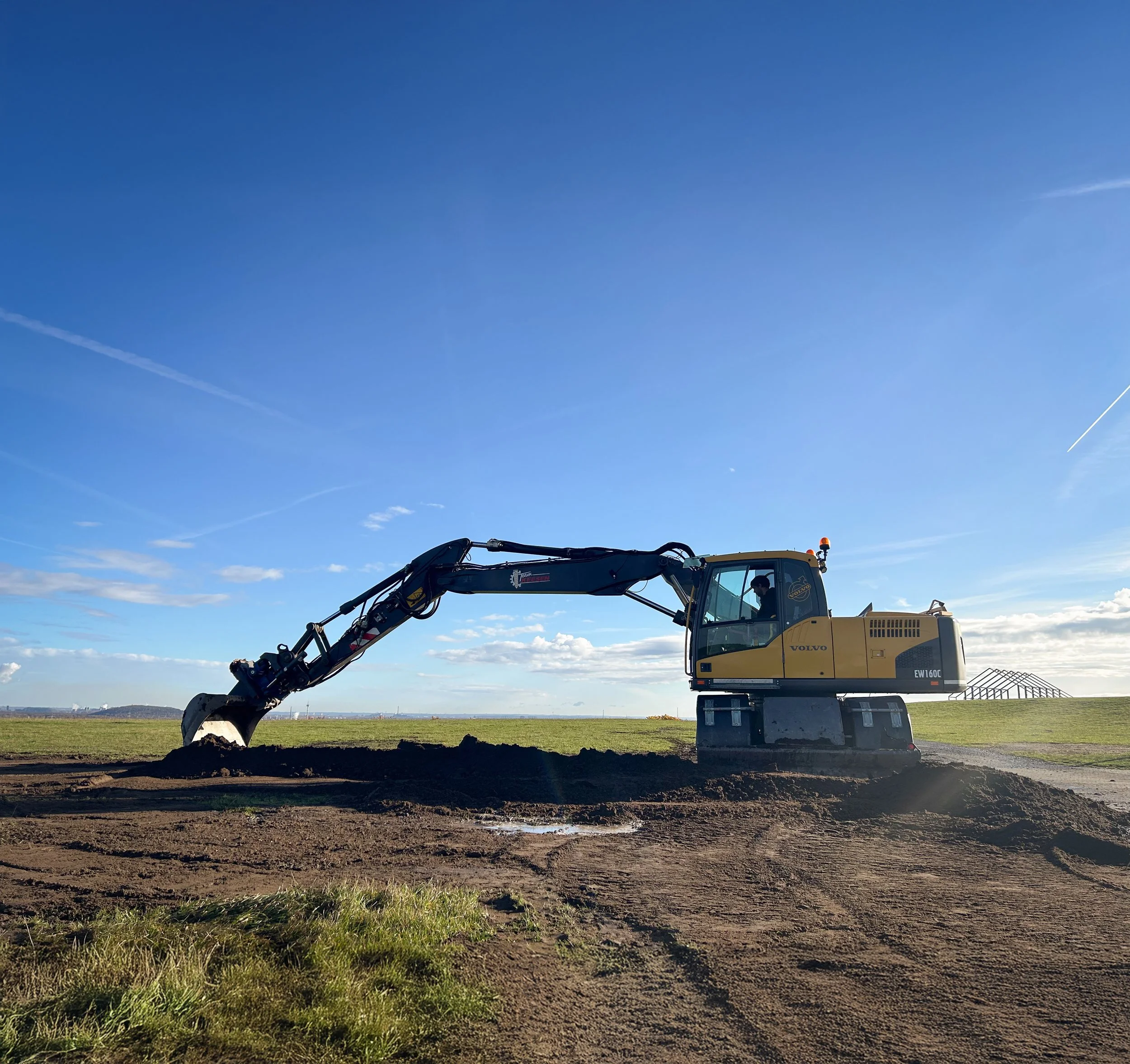 Ein gelber Volvo-Bagger bei der Erdarbeiten auf einem Feld unter blauem Himmel.
