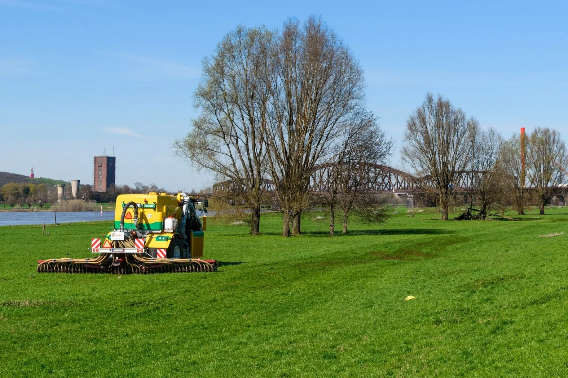 Ein landwirtschaftliches Traktor auf grünem Feld mit Bäumen, Fluss, Brücke und Gebäuden im Hintergrund an einem sonnigen Tag.