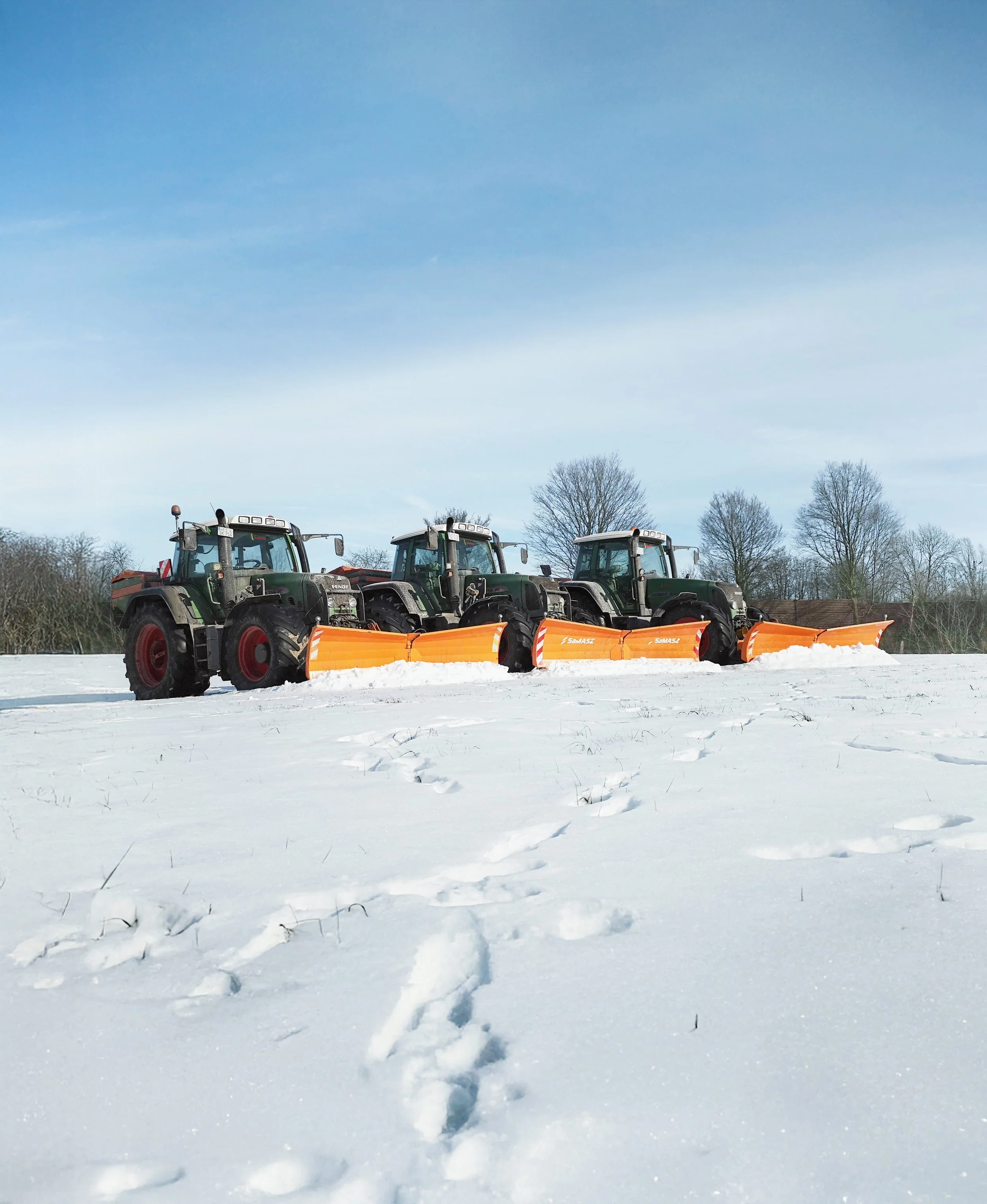 Drei landwirtschaftliche Traktoren mit Schneepflügen, die Schnee auf einem Feld entfernen, bei klarem Himmel.