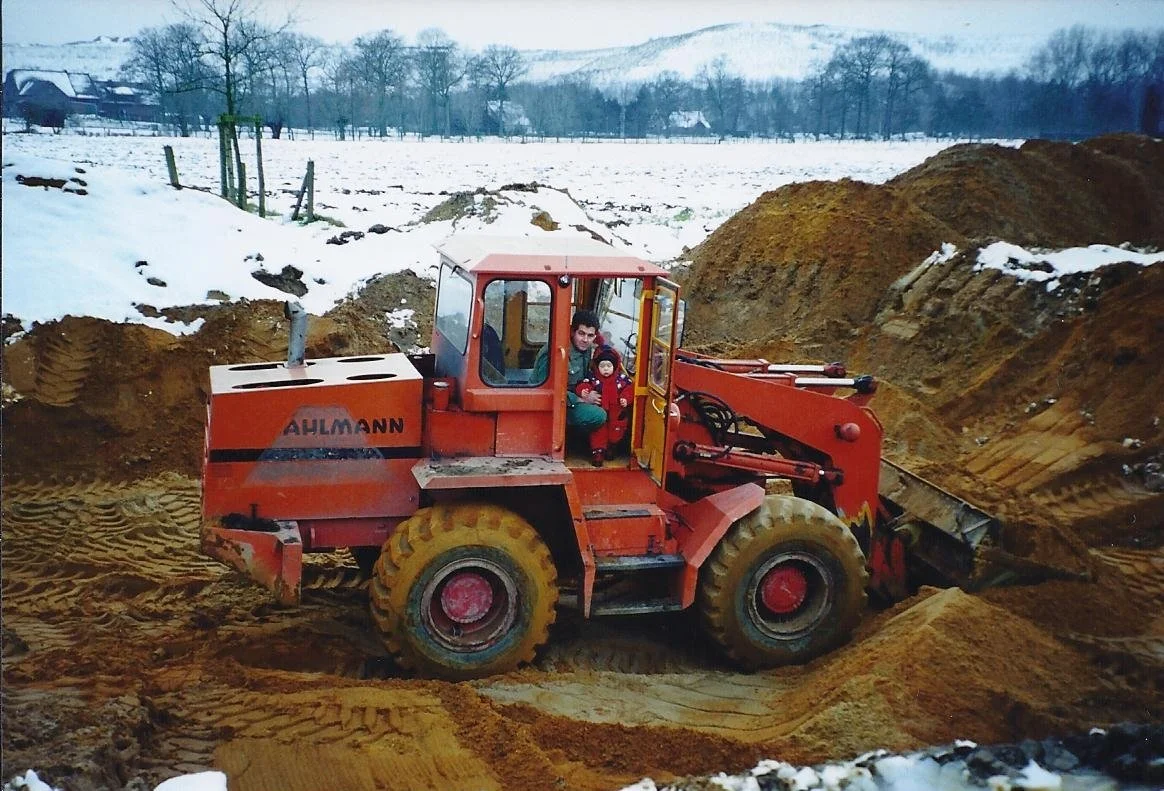 Ein roter Radlader arbeitet auf einer Baustelle im Schnee, während eine Person und ein Kind im Fahrerbereich sitzen.