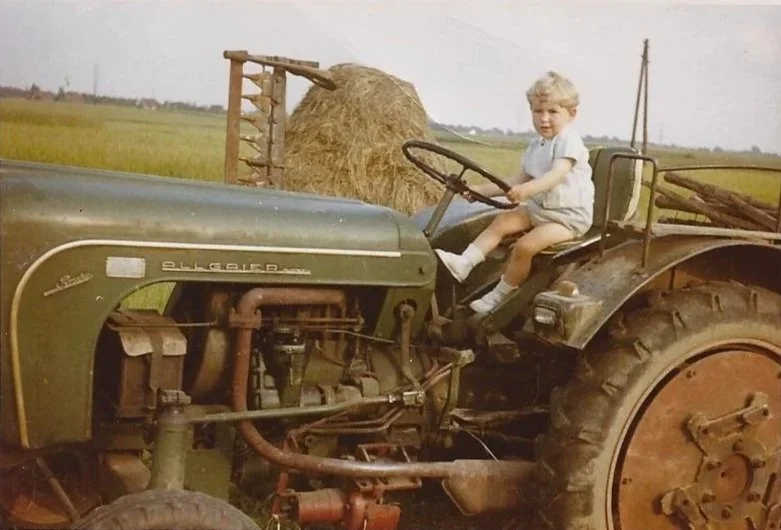 Ein Junge sitzt auf einem alten Traktor, mit einem Heuhaufen im Hintergrund auf einer landwirtschaftlichen Fläche.