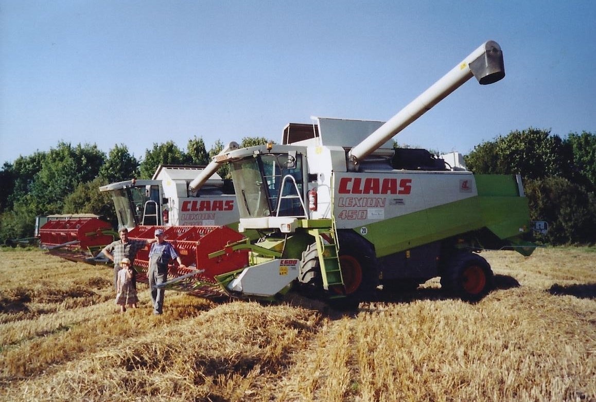 Eine Familie steht vor einer großen landwirtschaftlichen Maschine auf einer goldbraunen Getreidewiese, mit Bäumen im Hintergrund.