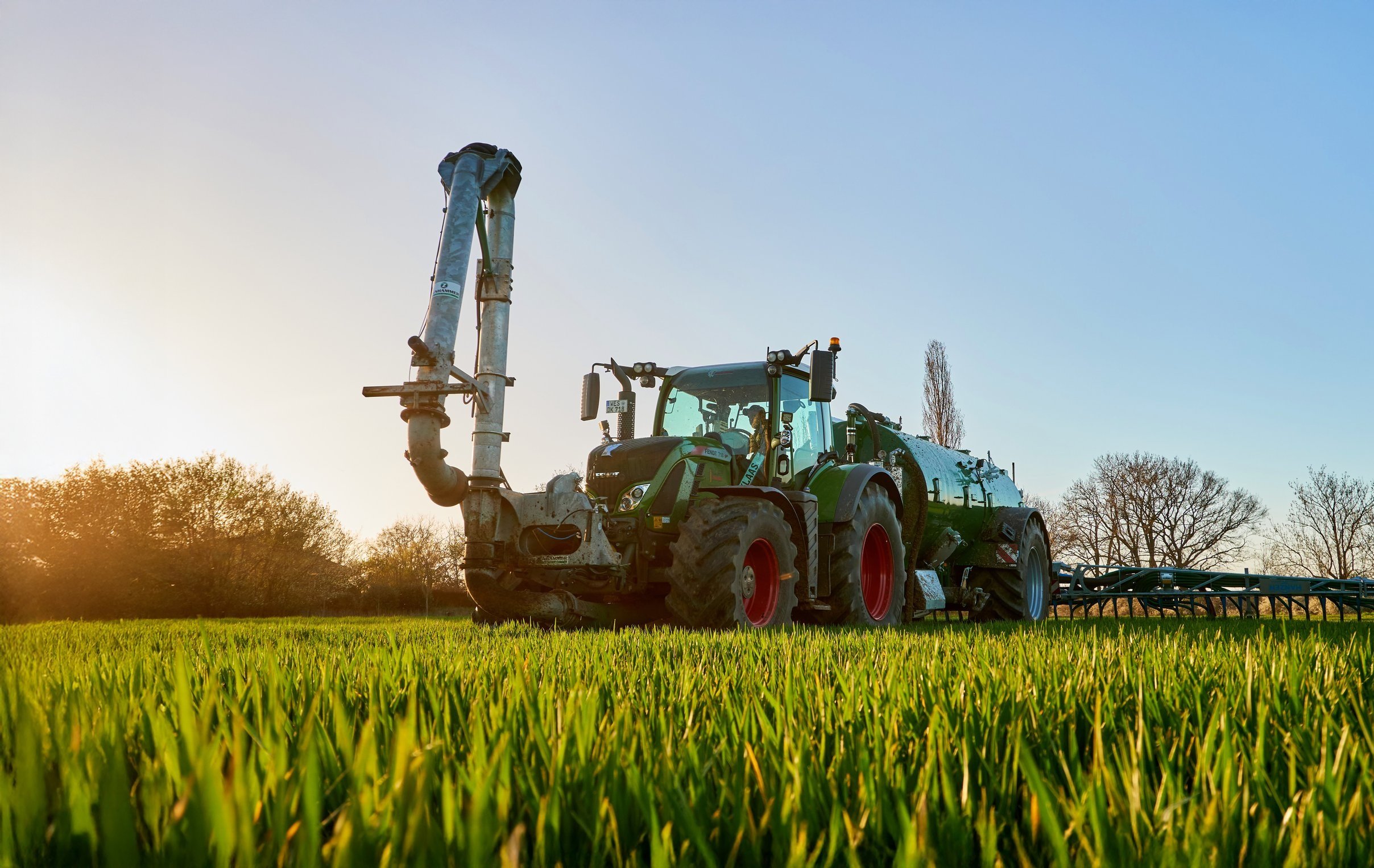 Grüner Traktor mit Sprühgerät im Feld, Sonnenaufgang im Hintergrund, Bäume im Hintergrund.