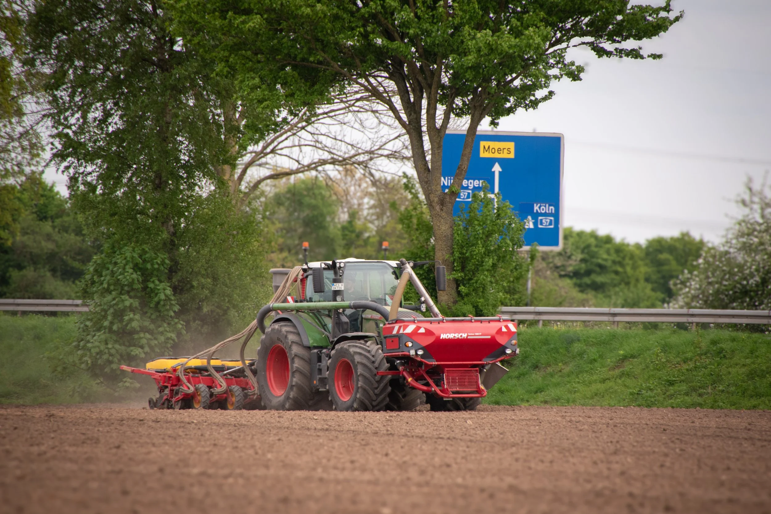 Traktor arbeitet auf einem Acker, hinter Bäumen und einem Verkehrszeichen in Deutschland.