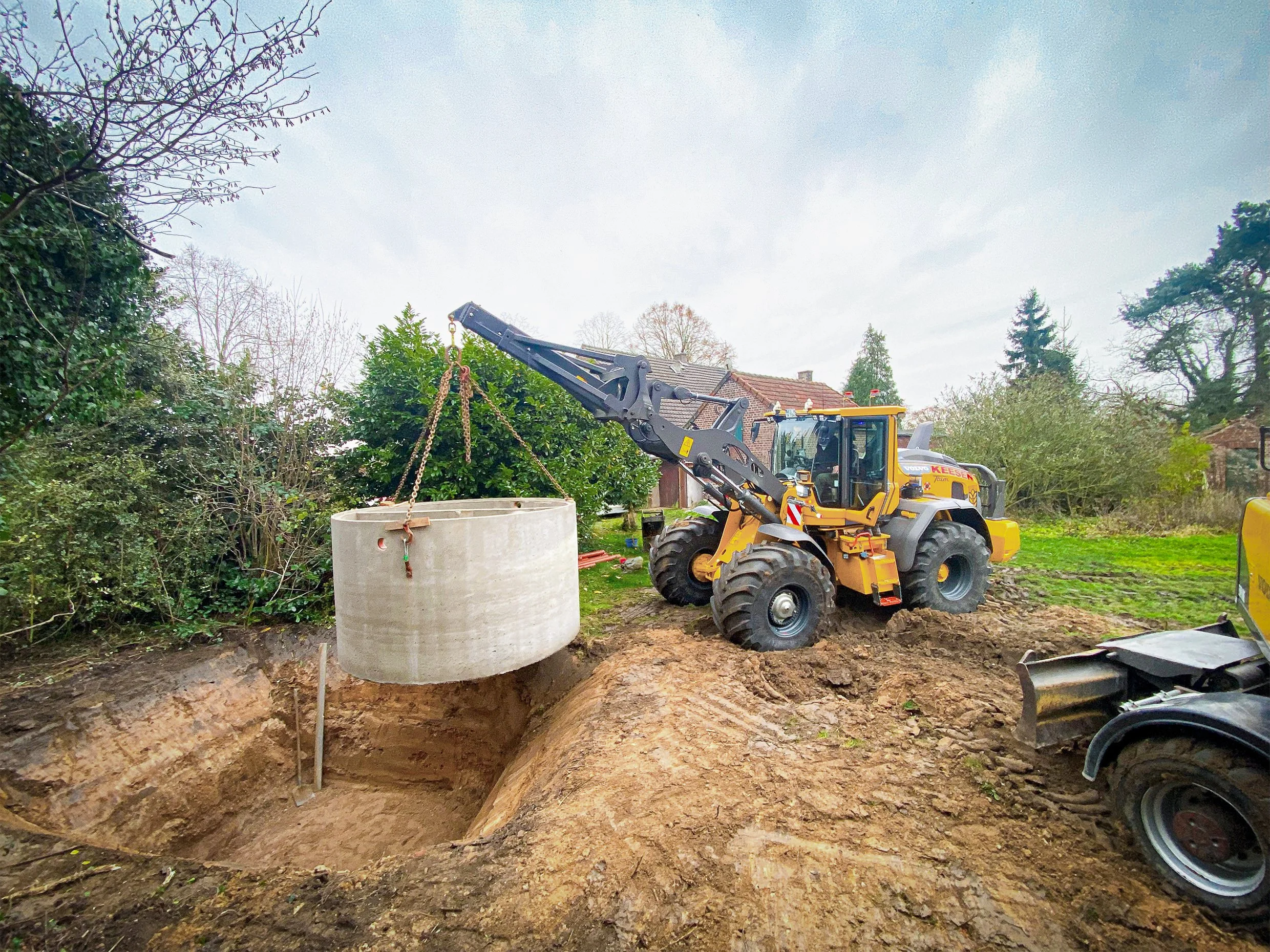 Bagger hebt einen runden Betontank aus einem Graben auf einer Baustelle in einem Garten mit Bäumen und Häusern im Hintergrund.