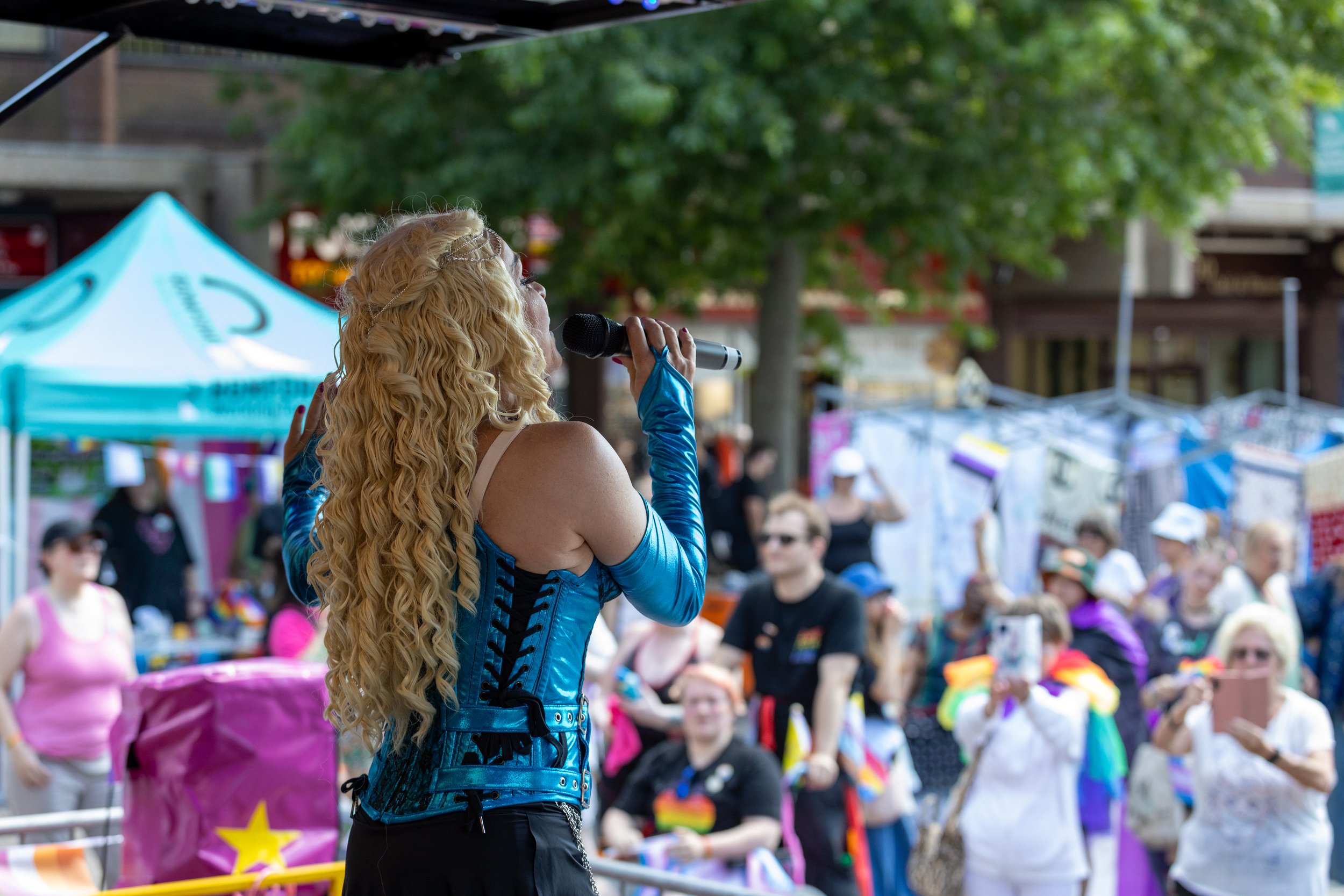 A performer with long, curly blonde hair singing into a microphone at an outdoor event, with a crowd of diverse people holding pride flags and taking photos in the background.