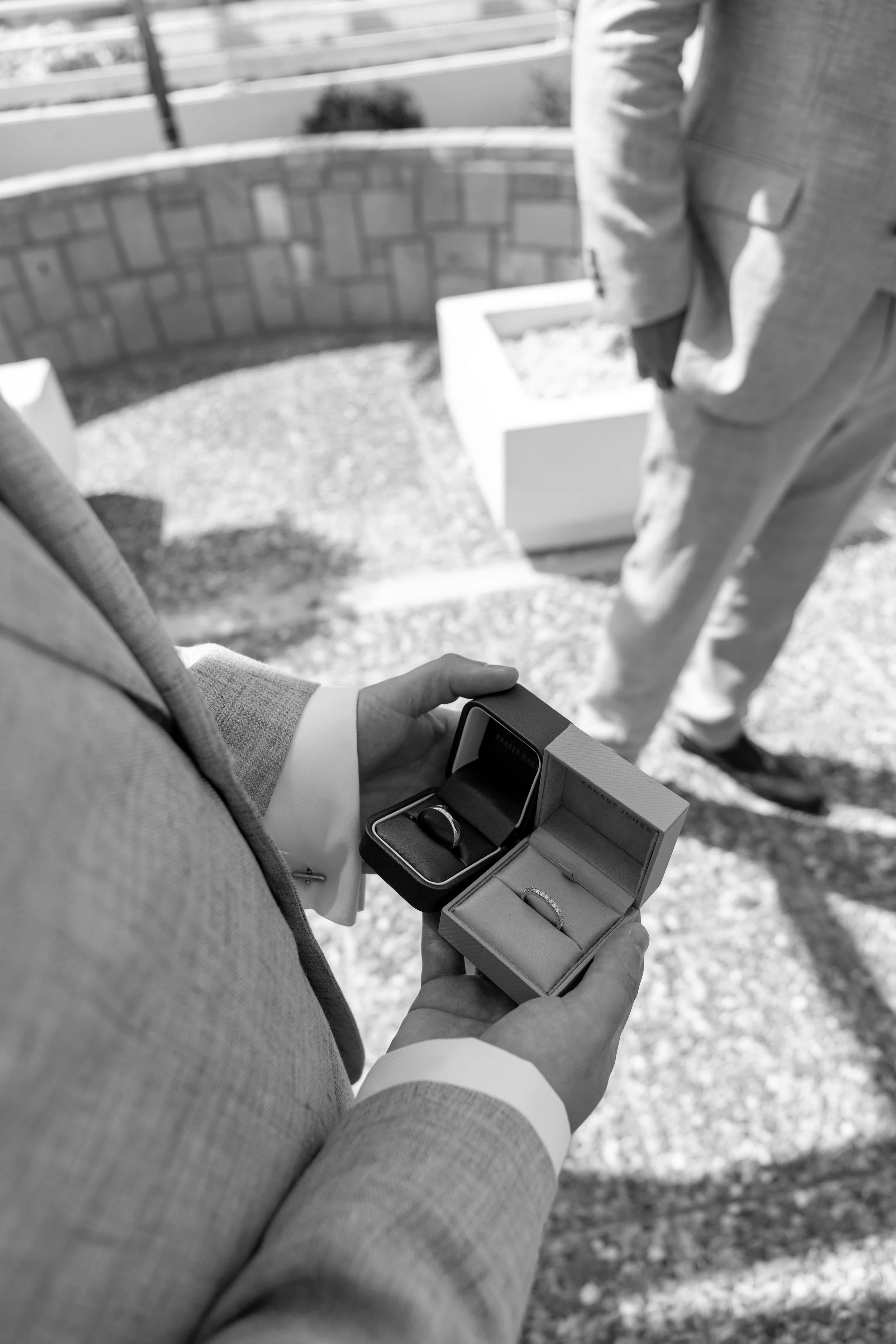 A man in a light suit is holding open a jewelry box displaying two rings, one in each box, in an outdoor setting with another person partially visible.