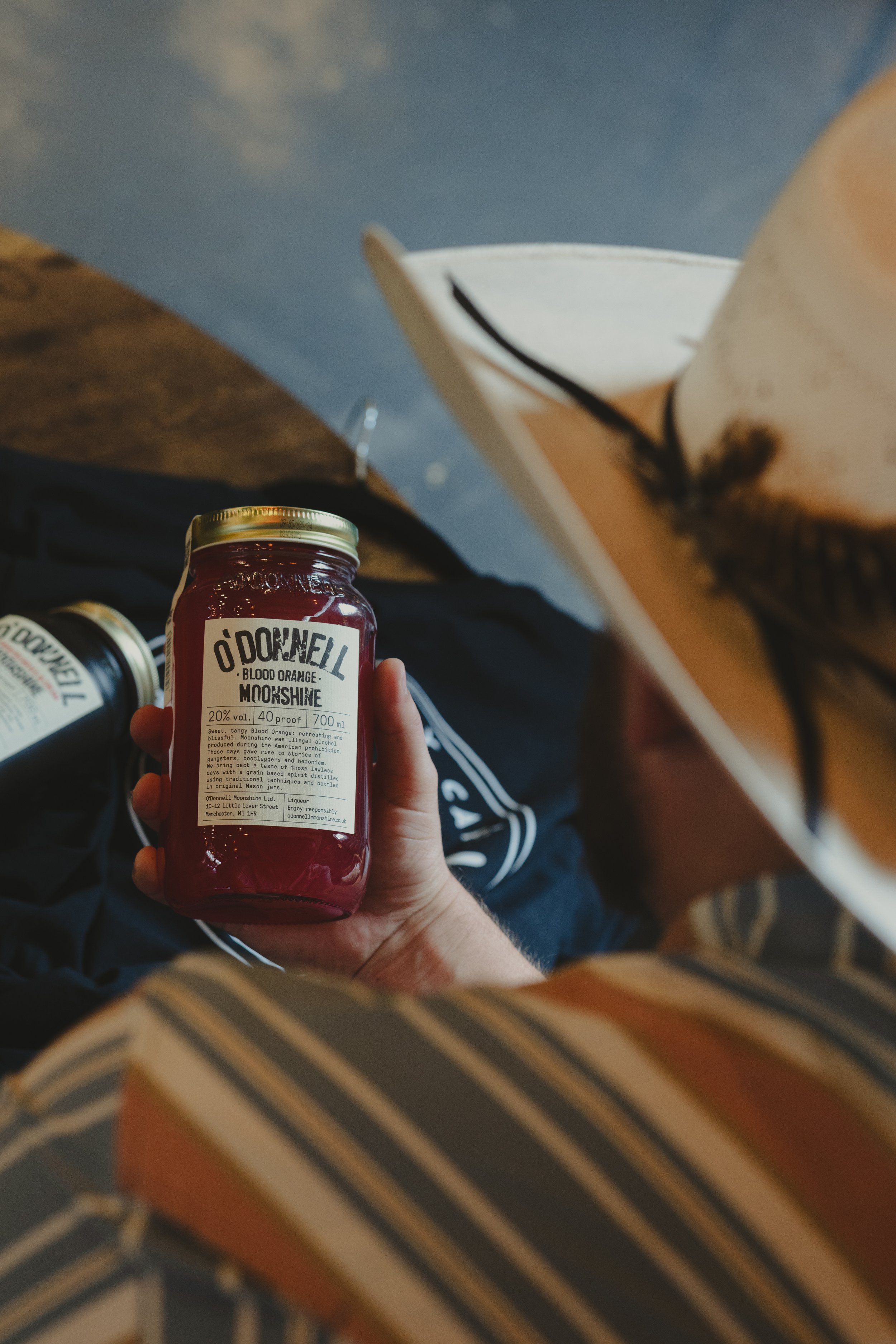 A person holding a jar of Downell Blood Orange Moonshine with a white label and gold lid, sitting on a dark cloth next to more jars, with a book partially visible on their lap.