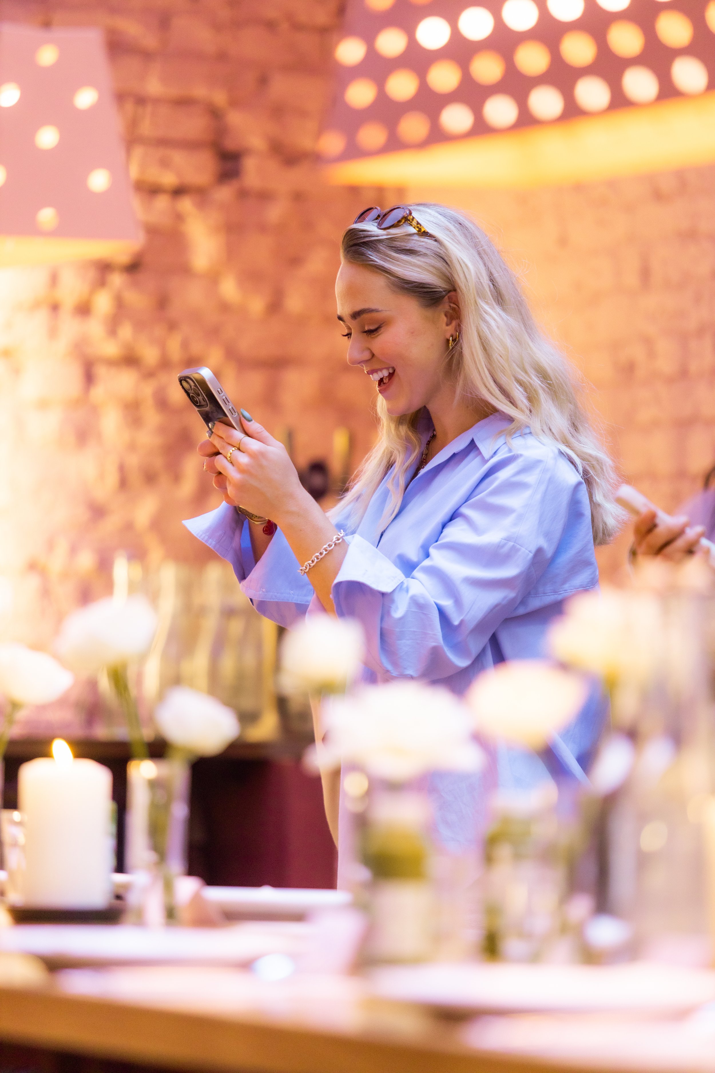 A woman with long blonde hair wearing a light blue shirt, smiling as she looks at her phone in a warmly lit setting with candles and flowers on a table in front of her.