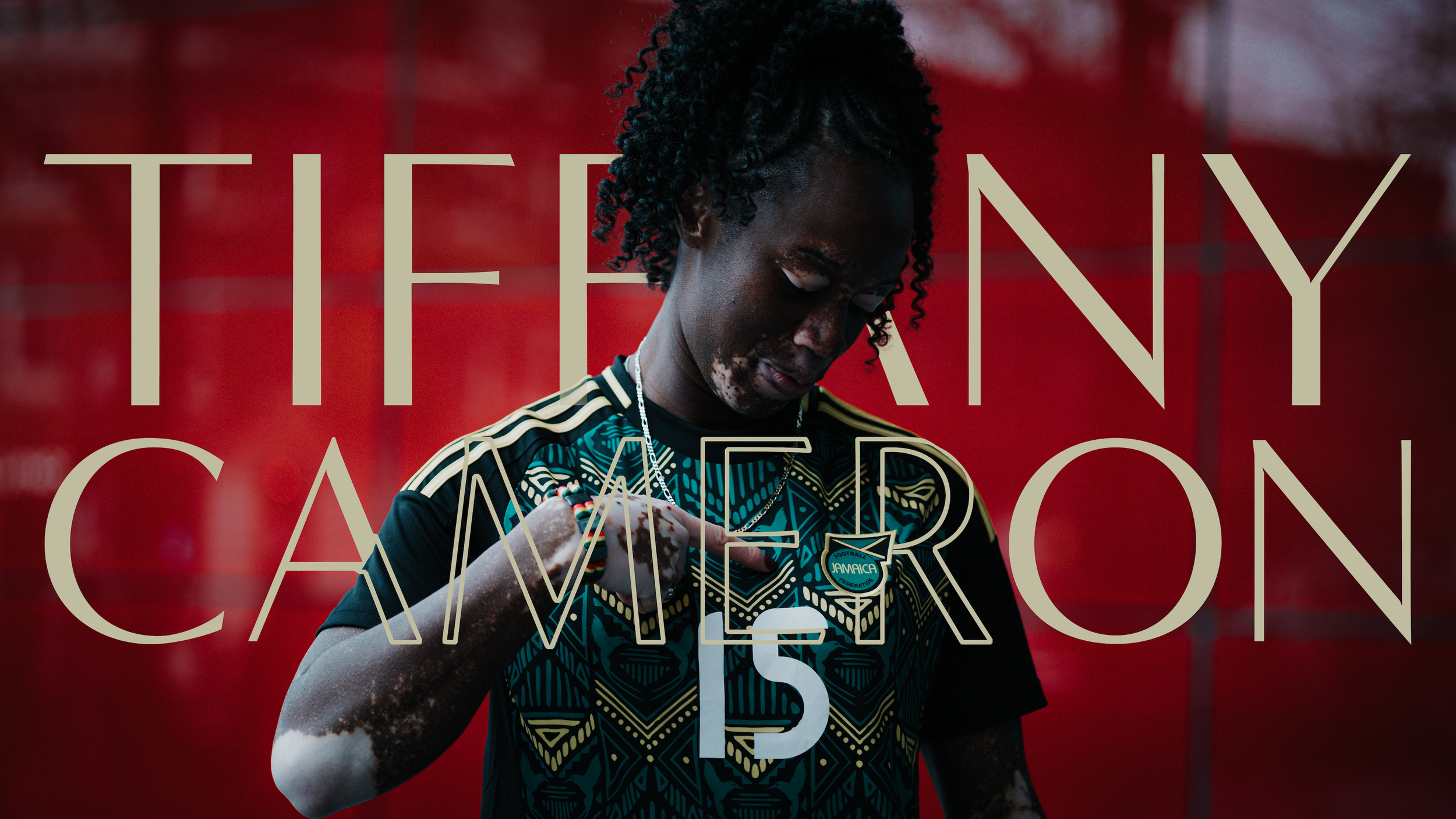 A young woman with dark skin and curly hair wearing a black and turquoise Jamaican football jersey, standing against a red background, looking down while holding a bracelet with both hands. Overlaid text reads 'Tiffany Cameroon 15'.