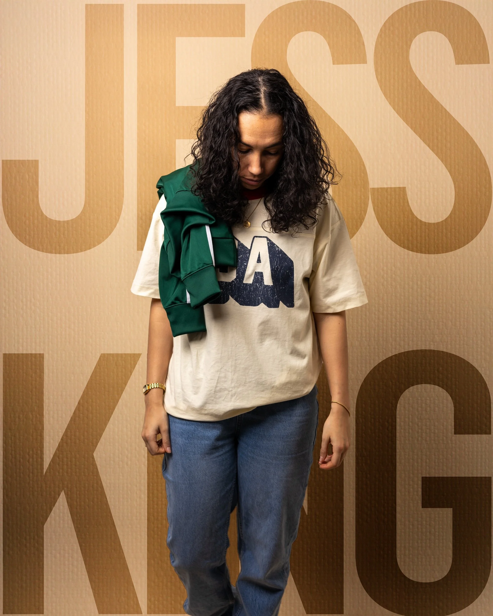 A woman with curly dark hair, wearing a beige T-shirt and jeans, is looking down. She has a green jacket draped over her shoulder and is standing in front of a beige background with large, faint text.