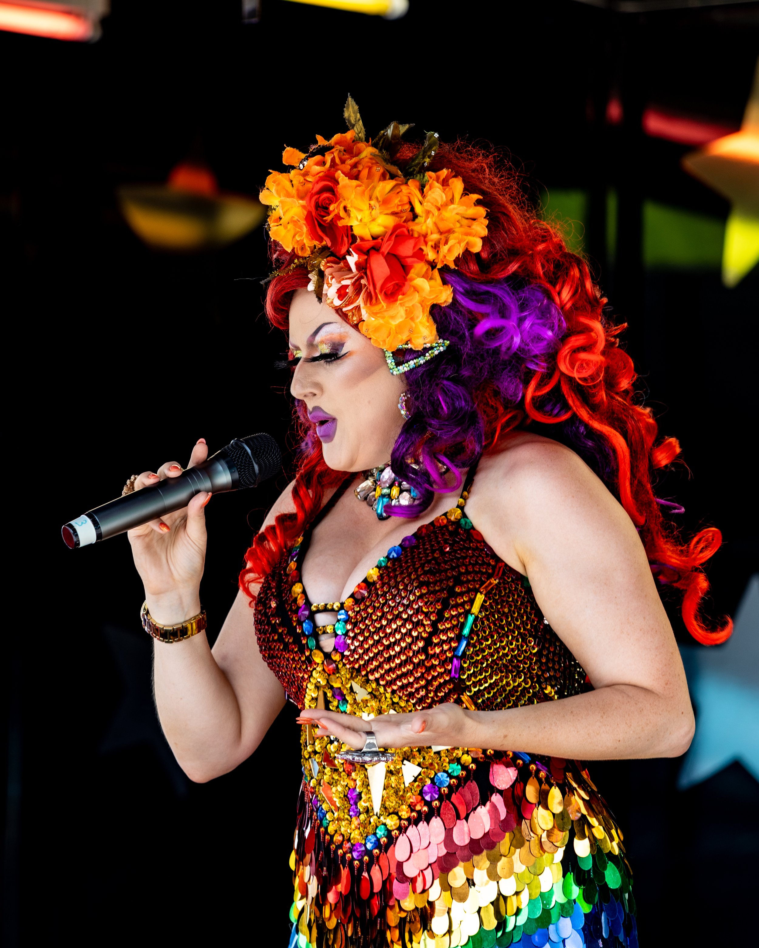 Performer singing on stage, wearing a colorful, sequined dress with rainbow colors and a large floral headdress with orange, yellow, and red flowers, and purple and red curly hair.