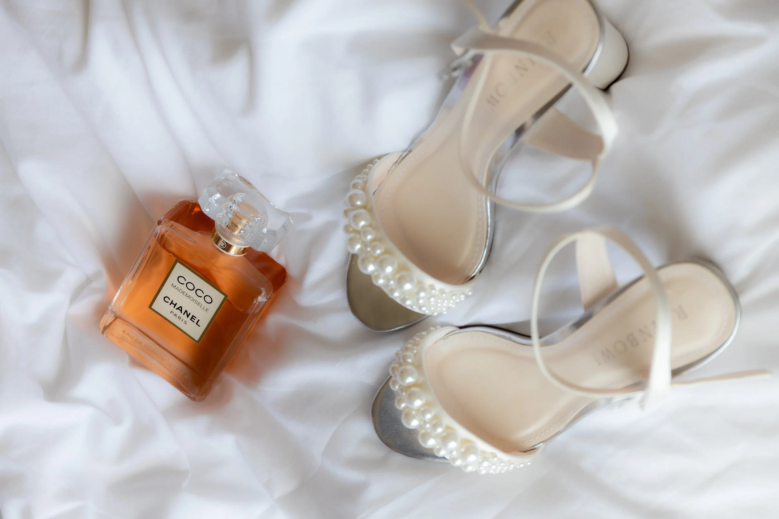 A bottle of Chanel Coco Mademoiselle perfume and a pair of high-heeled shoes with pearl embellishments on a white satin fabric.