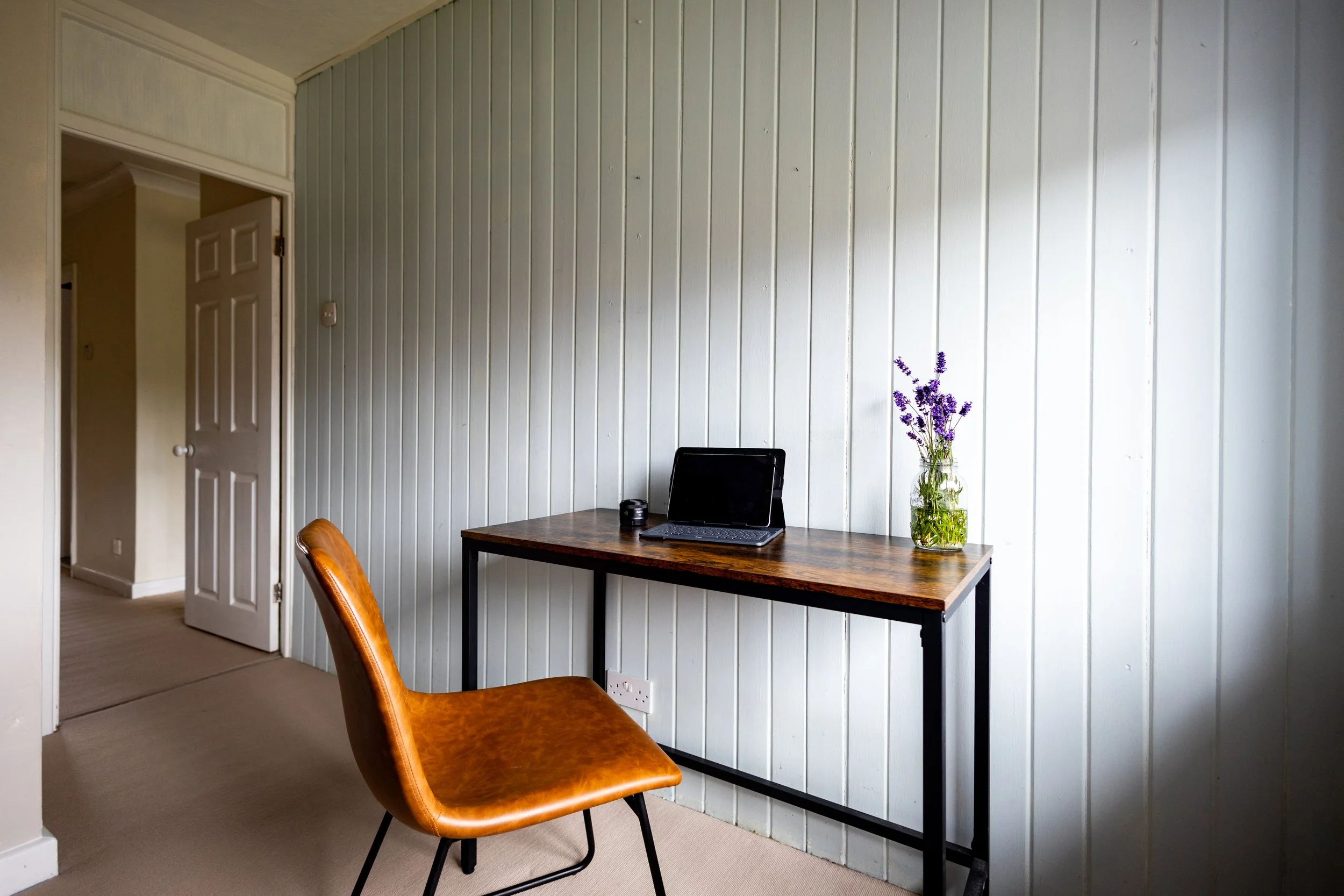 A small workspace with a wooden desk, a brown leather chair, a laptop, and a vase of purple flowers on the desk in a room with white panel walls.