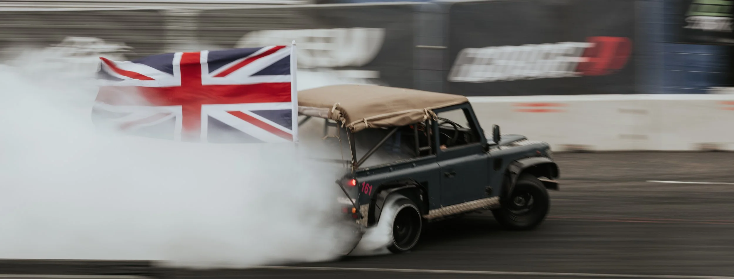 A dark-colored off-road vehicle with a beige soft top driving on a race track, emitting smoke from the rear tires, with a UK flag attached to the back of the vehicle.