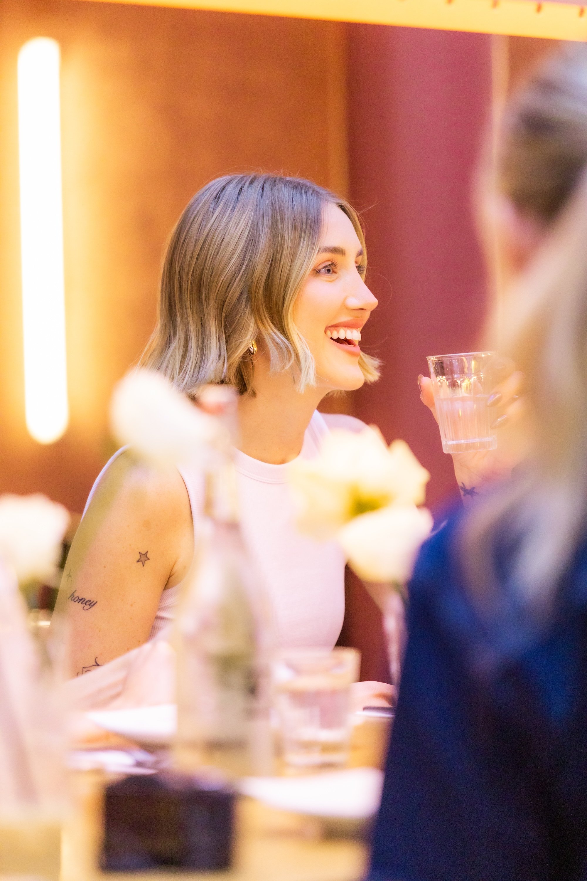 A young woman with shoulder-length blonde hair, smiling and holding a glass of water, sitting at a table during a social gathering or party.