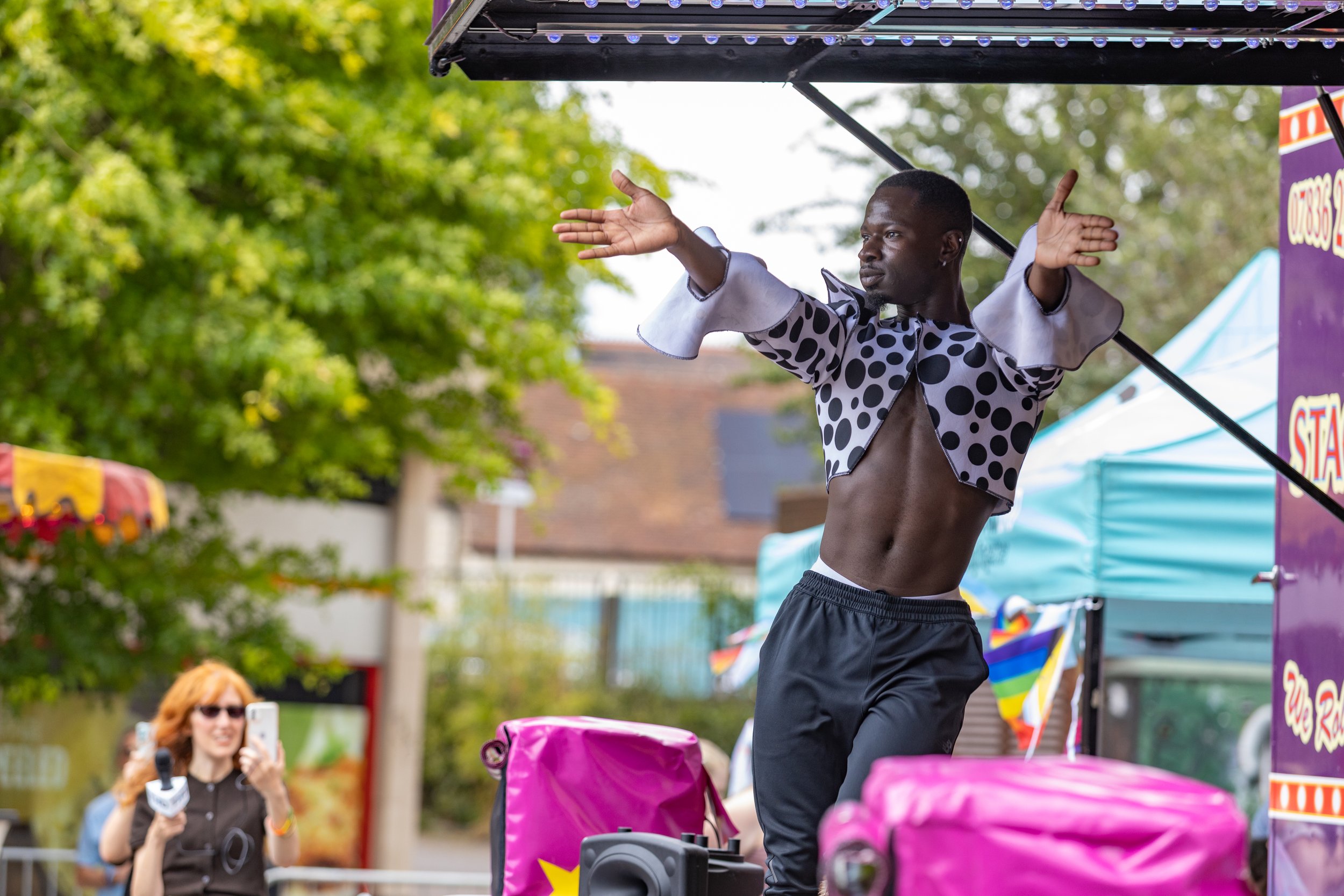 A man performing on an outdoor stage, wearing a polka-dot jacket with open chest showing muscular abdomen, raising his arms in a dramatic gesture. A woman with red hair and sunglasses is in the background, recording with her phone.