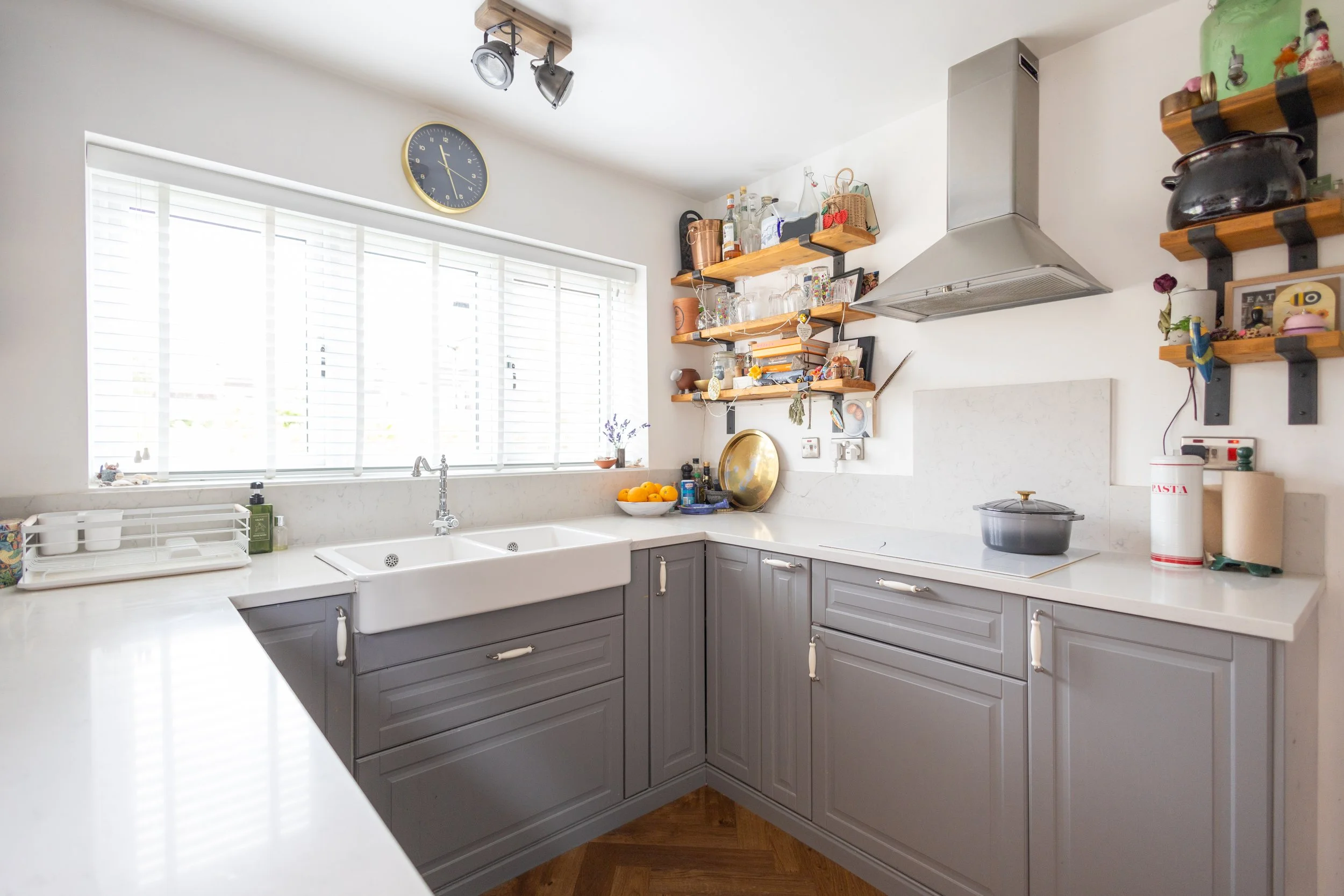 Kitchen with gray cabinets, white countertop, white sink, large window with blinds, wall-mounted wooden shelves holding bottles, jars, decorations, a range hood, and a bowl of oranges.
