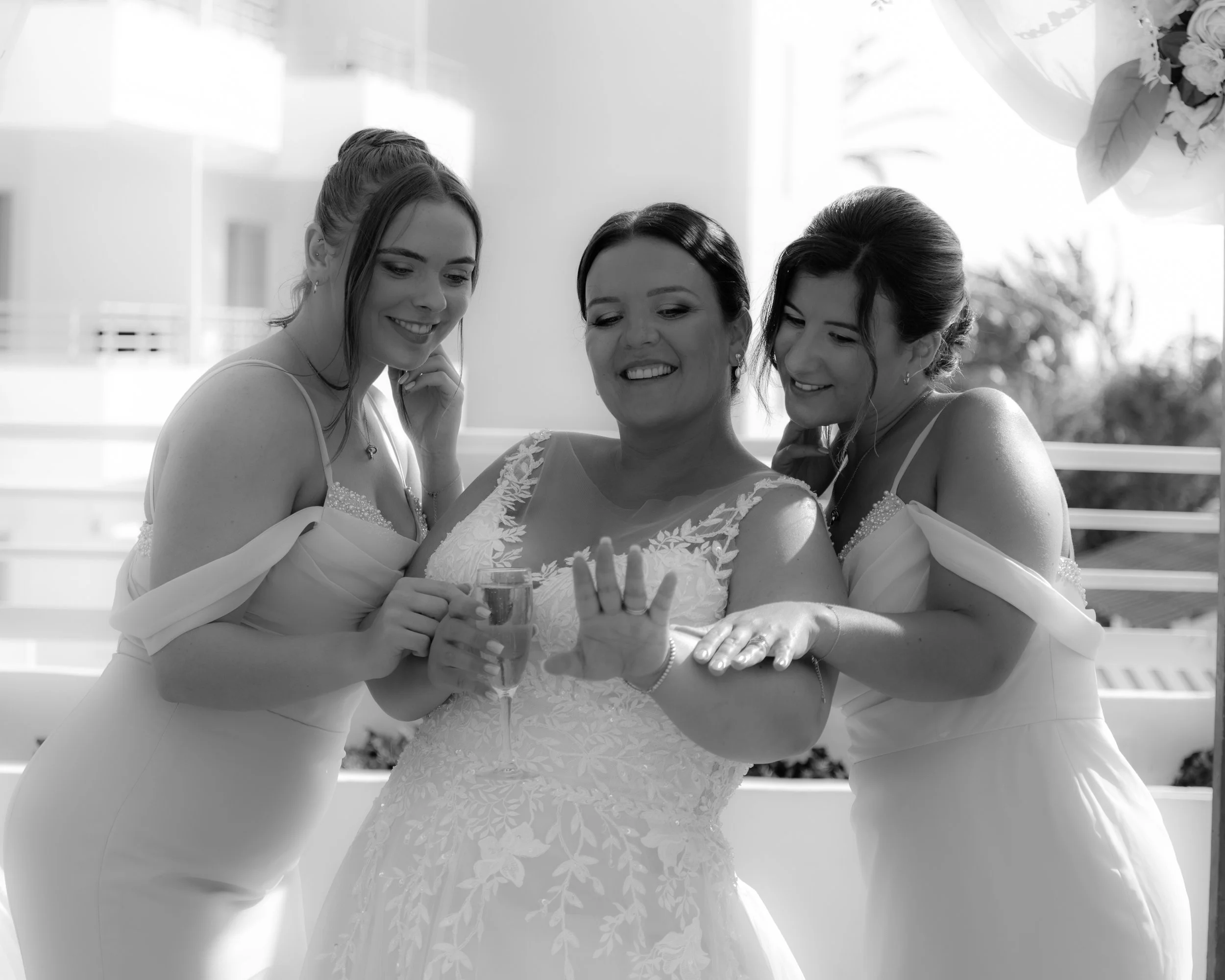 Three women dressed in white dresses, smiling and showing a first look at a wedding, with one woman holding a glass in her right hand, the middle woman has her left hand raised showing a ring, and the woman on the right is touching the middle woman's