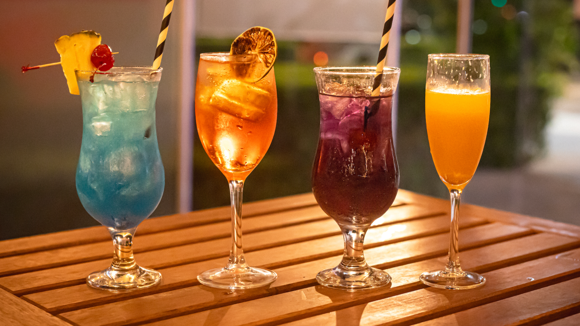 Four colorful cocktails on a wooden table at dusk, garnished with fruits and straws.