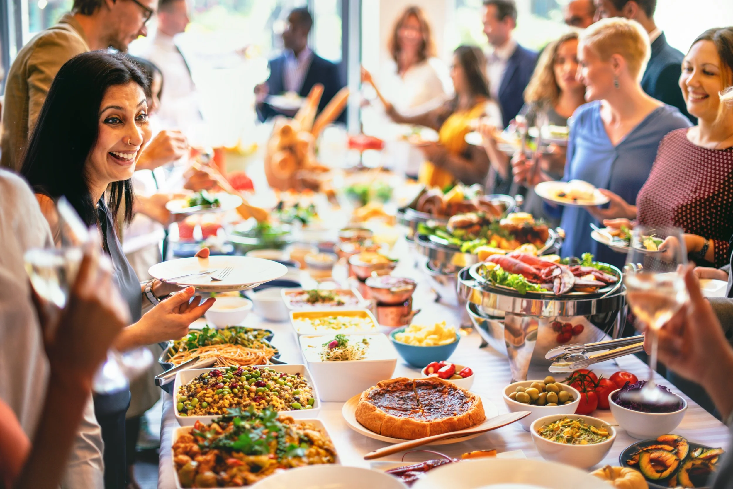 A diverse group of people enjoying a buffet-style meal at a social gathering, with various dishes and drinks on the table.