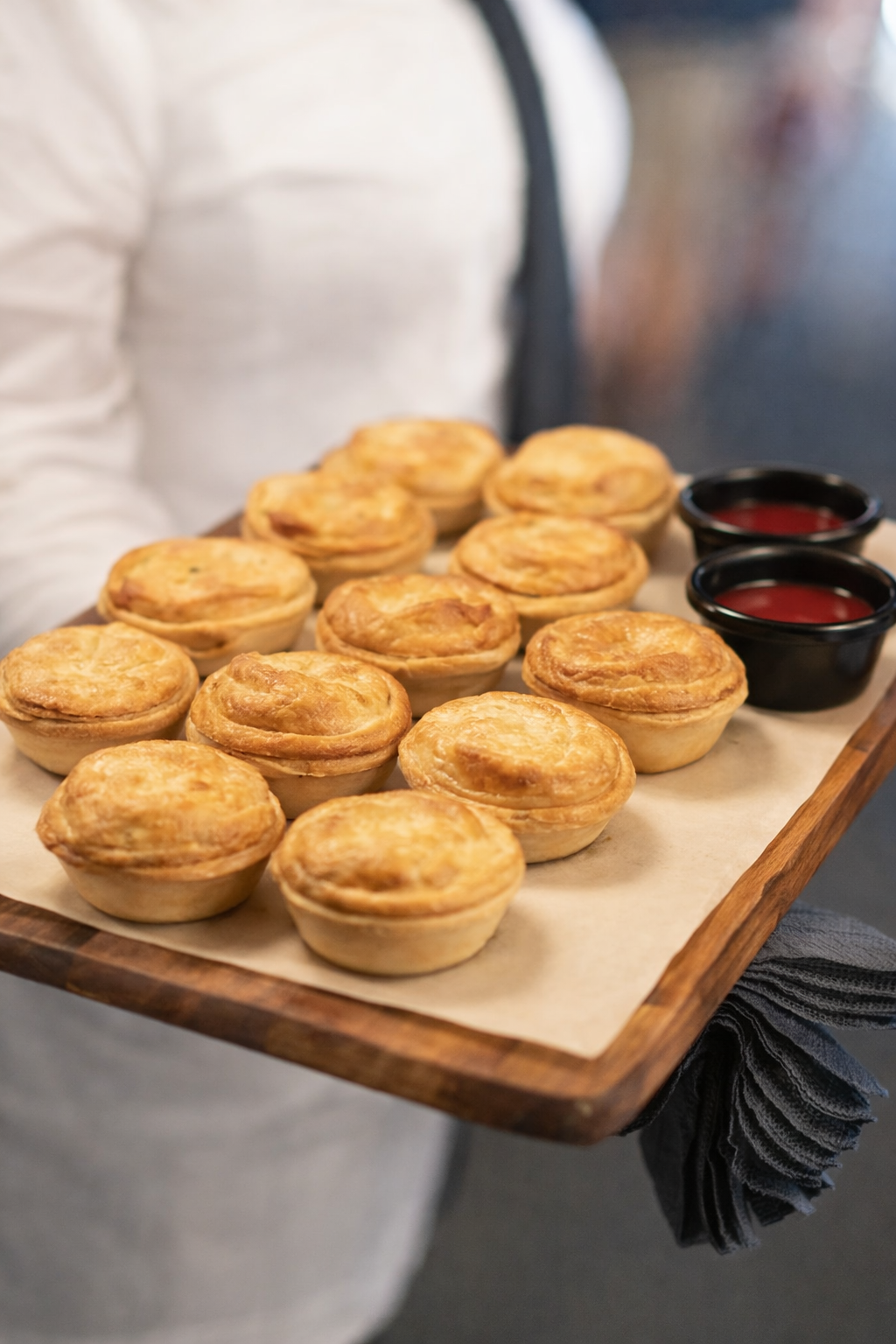 A person holding a wooden tray with 14 chicken pot pies and two small bowls of dipping sauce.