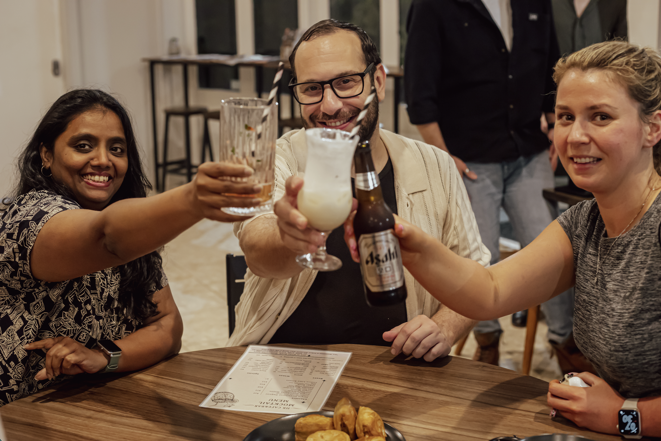 Three friends at a restaurant clinking drinks and smiling at the camera, with a menu and plate of snacks on the table.
